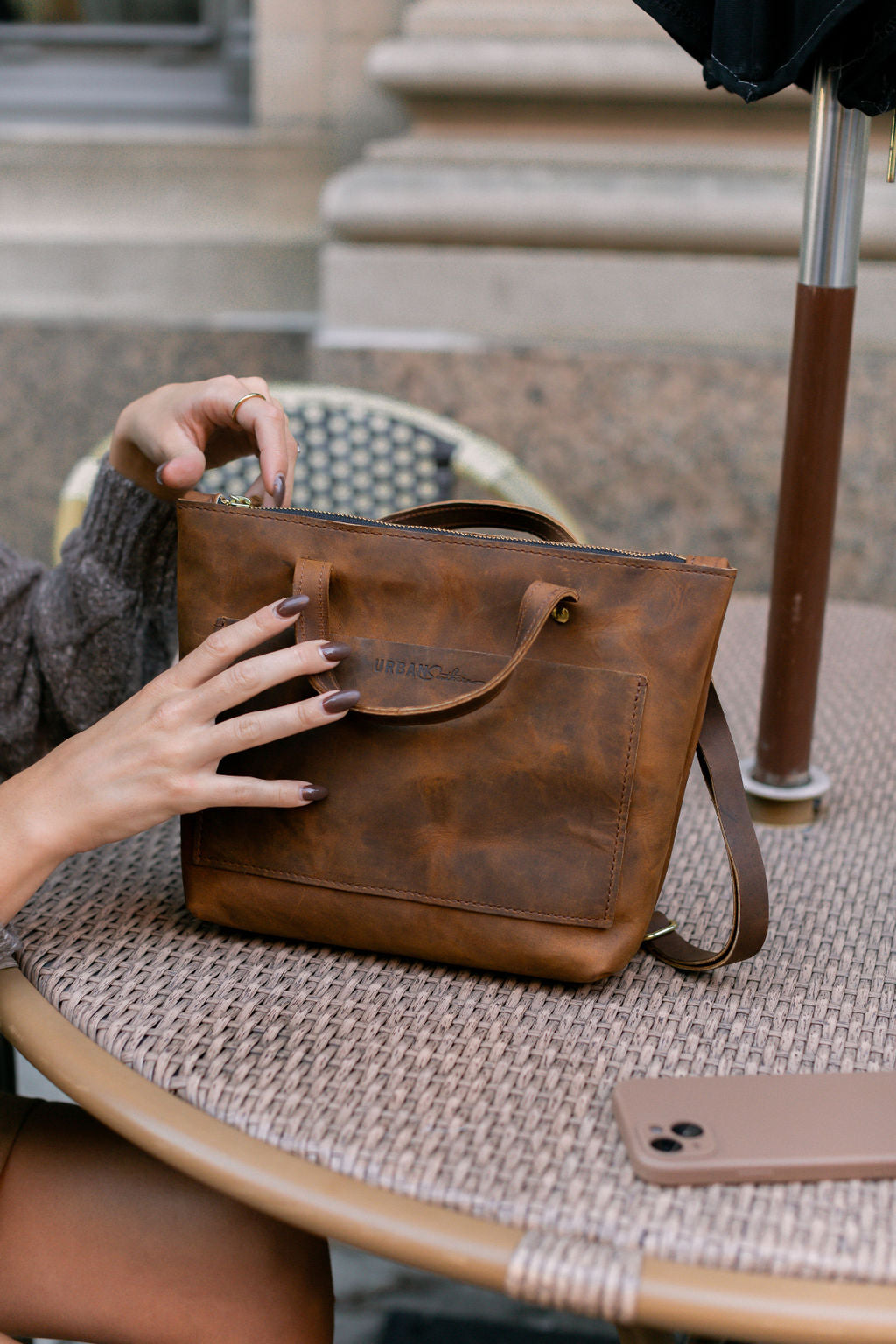 A woman sitting at a cafe table, begins unzipping a rustic brown Quinn Crossbody.