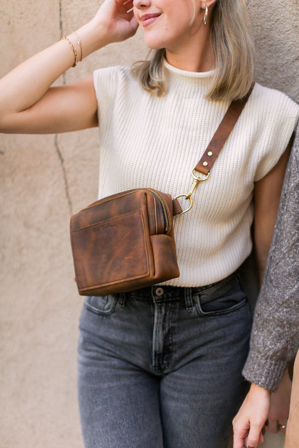 A woman stands next to a beige wall, on her chest she is wearing a rustic brown Urban Sling.
