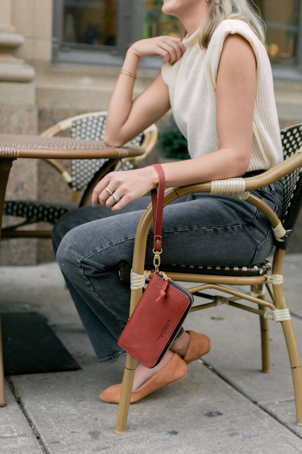 A woman sitting at an outdoor cafe, sits in a chair, while a merlot Urban Zipper Wallet hangs down from her wrist.
