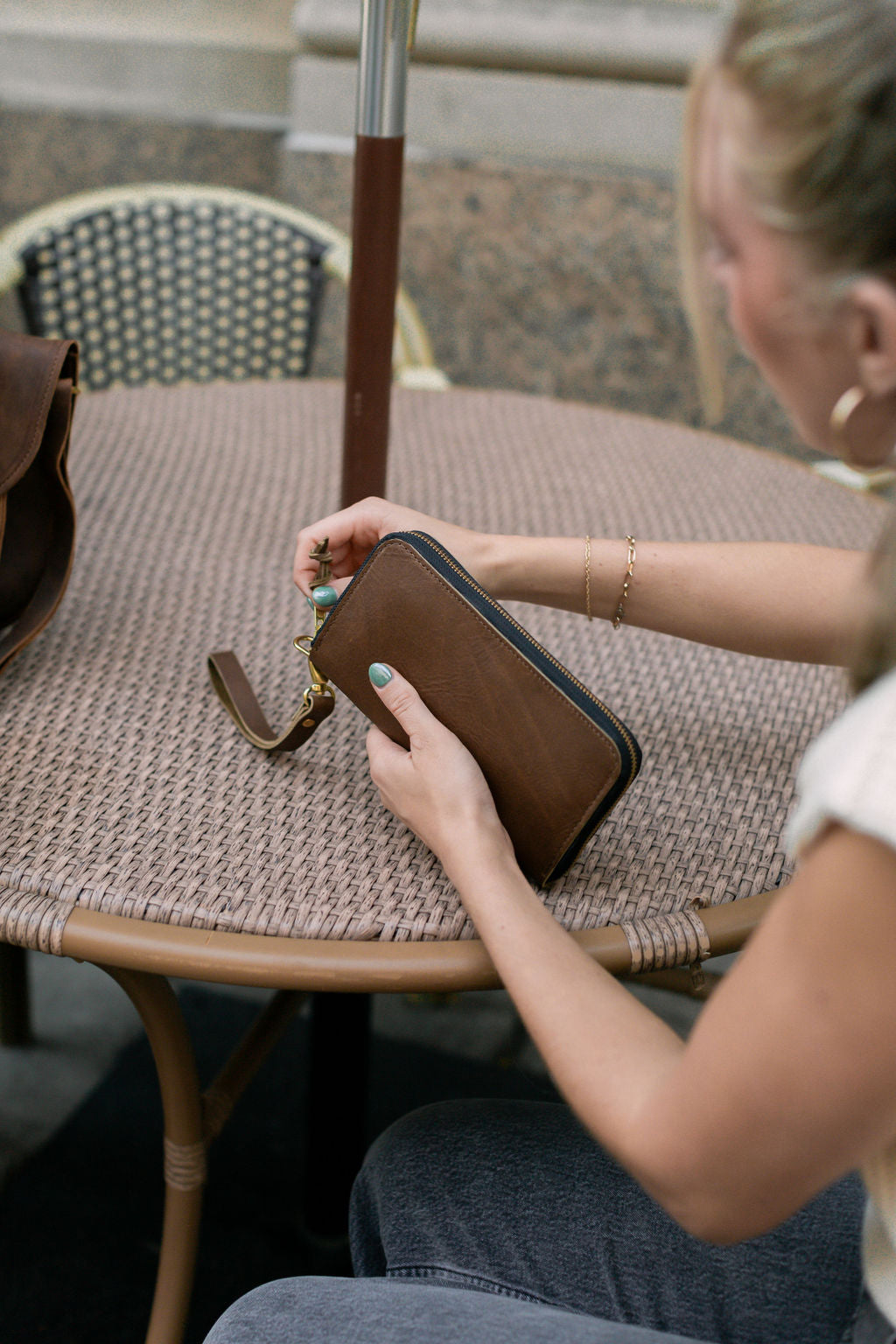 A woman sitting at an outdoor cafe holds and begins unzipping a mocha Urban Zipper Wallet.