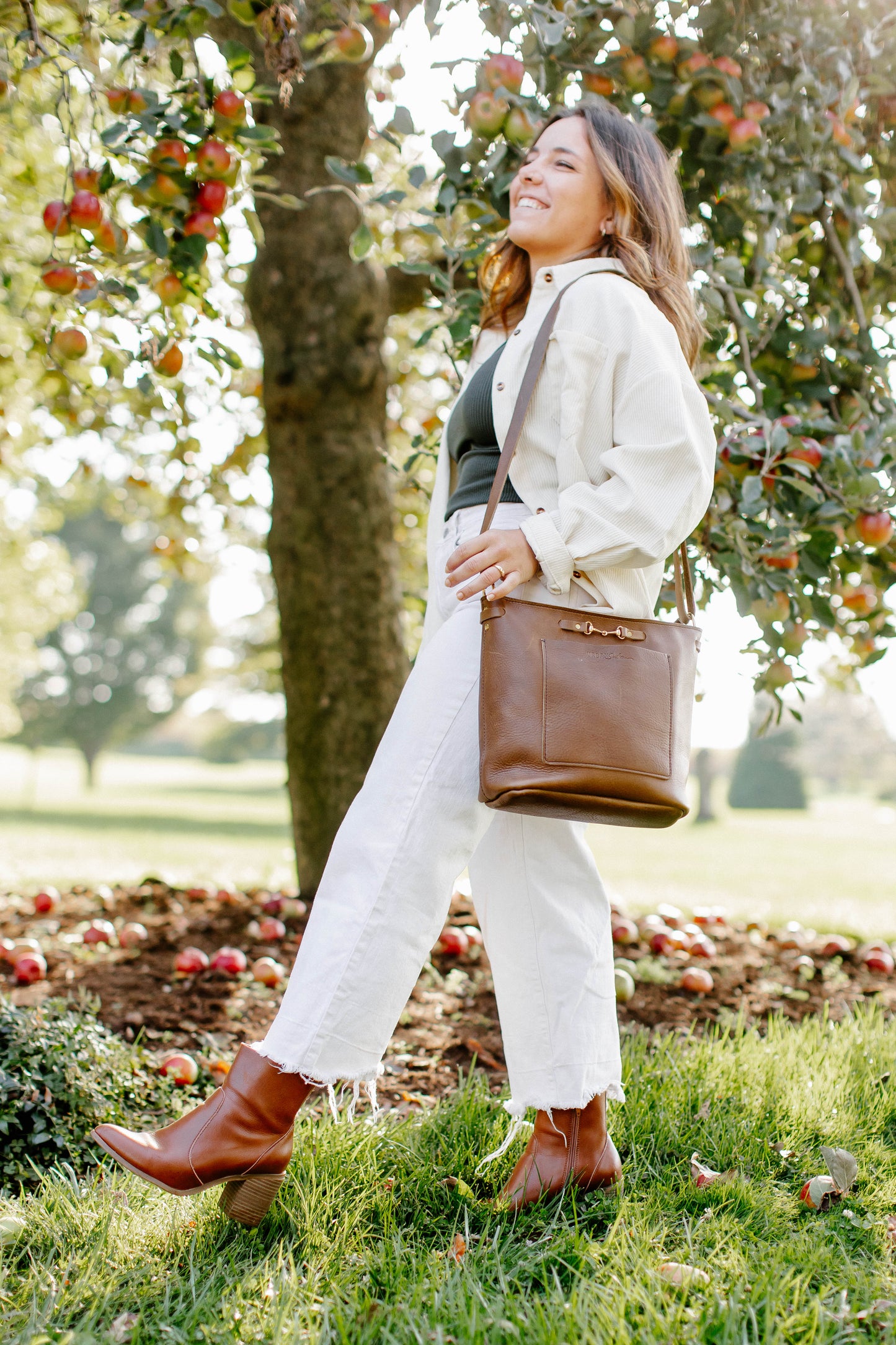 A woman wearing a mocha Lexington Julep leather handbag on her shoulder, walks through an apple orchard.