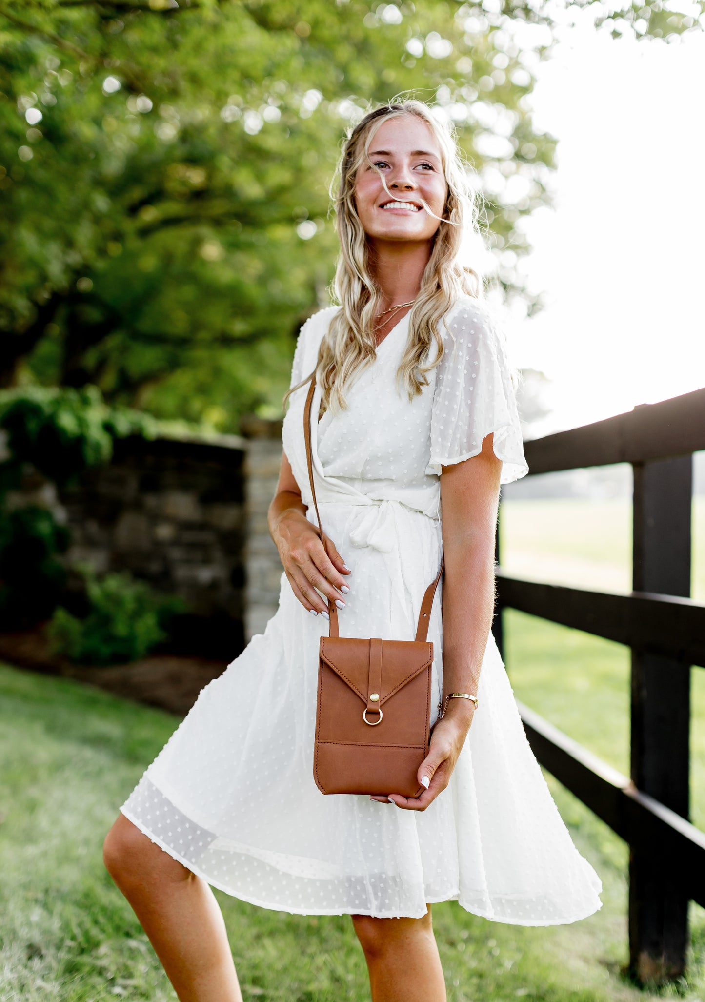 A woman wears a honey Postal Crossbody across her body, while showcasing the details of this leather bag.