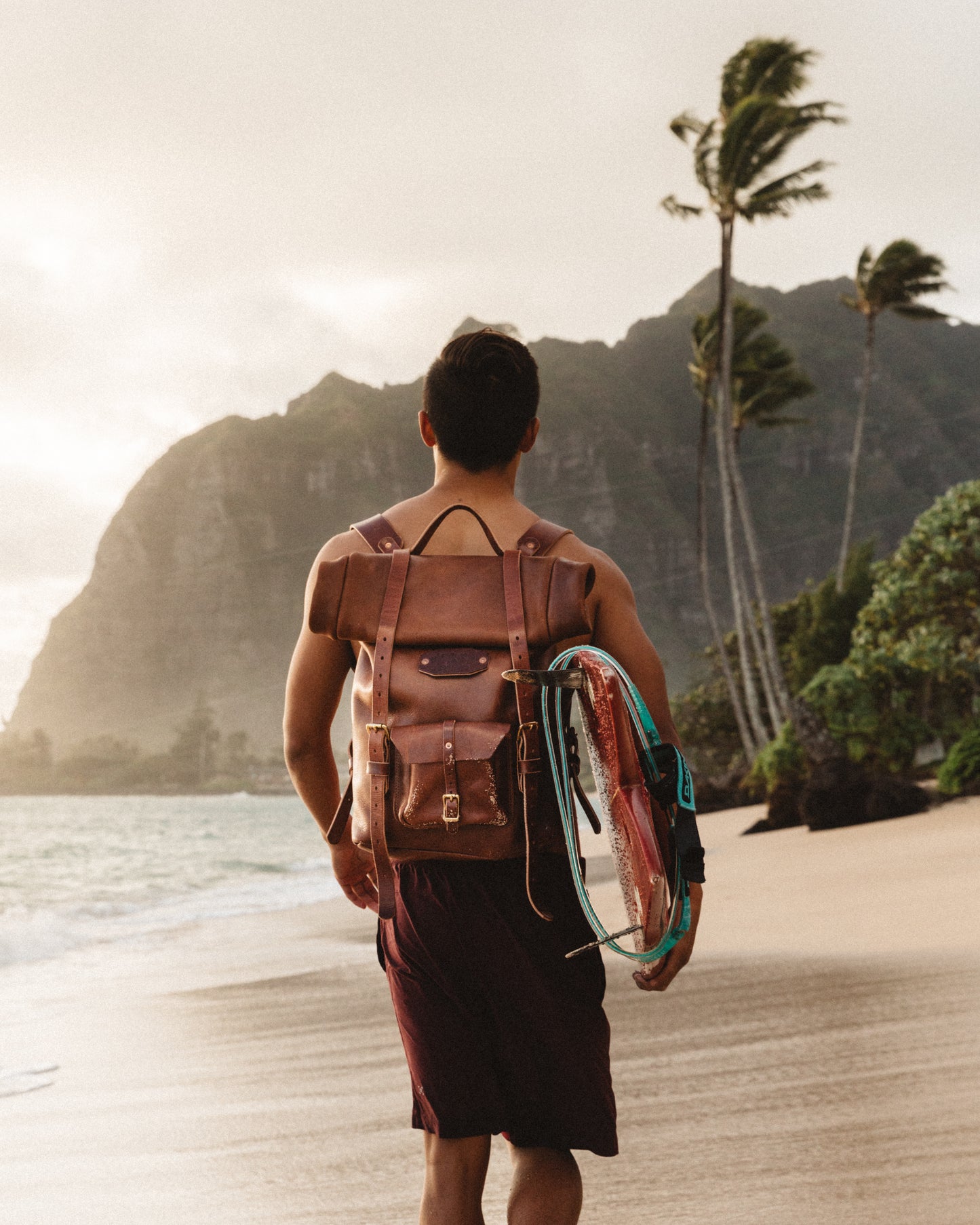 Man wearing a brown leather backpack holding a surfboard walking along a beach with palm trees and mountains in the background.