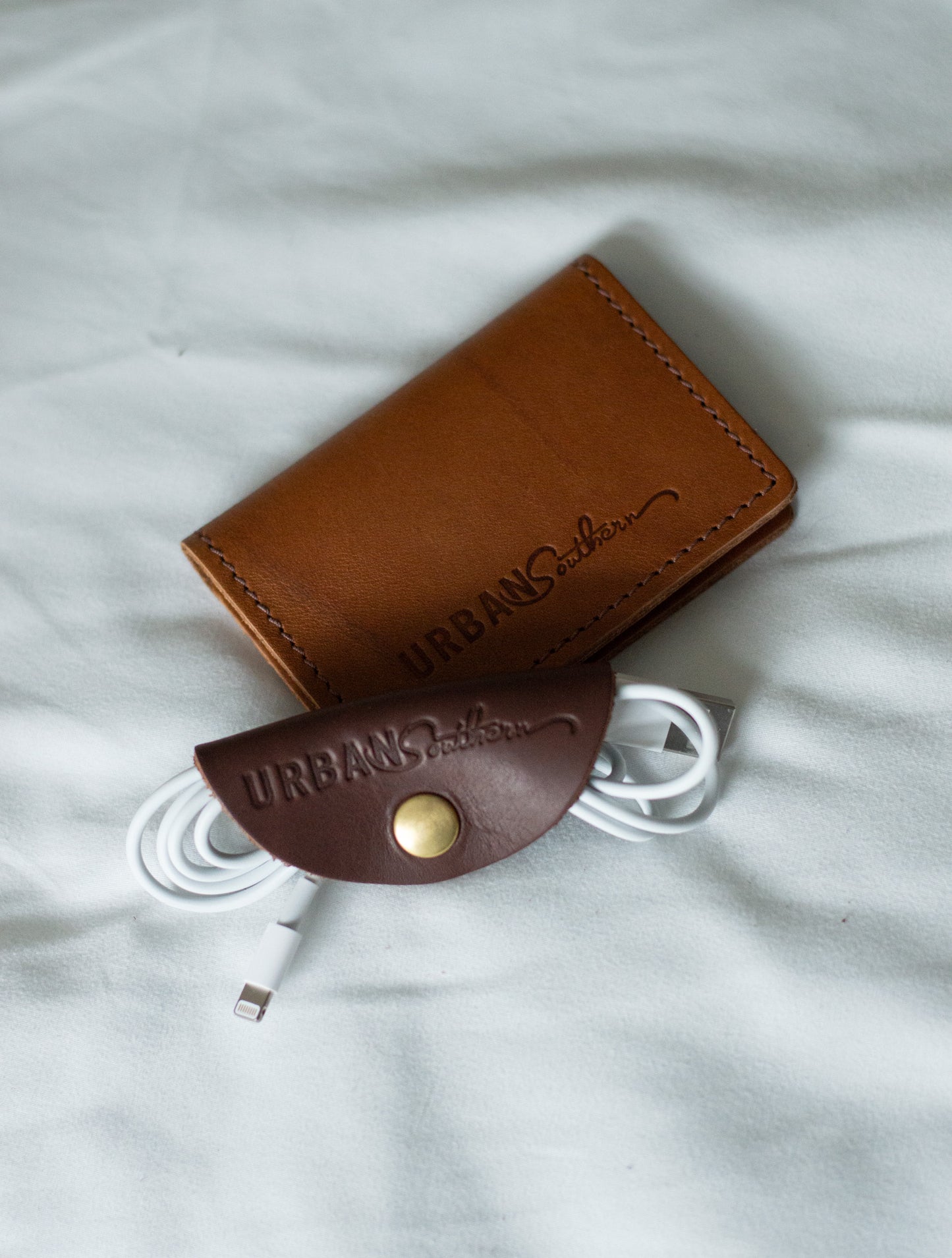 A chestnut leather Urban Cord Wrap neatly organizes a charging cable, while laying next to it is a honey Urban Card Wallet.