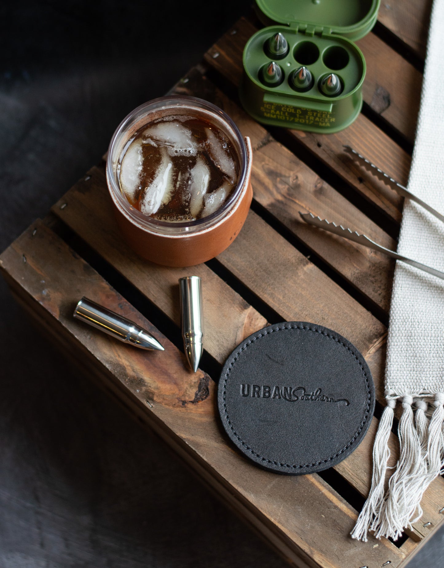 A Black, Urban Southern branded coaster sits on top of a wooden crate with a glass of whiskey next to it.