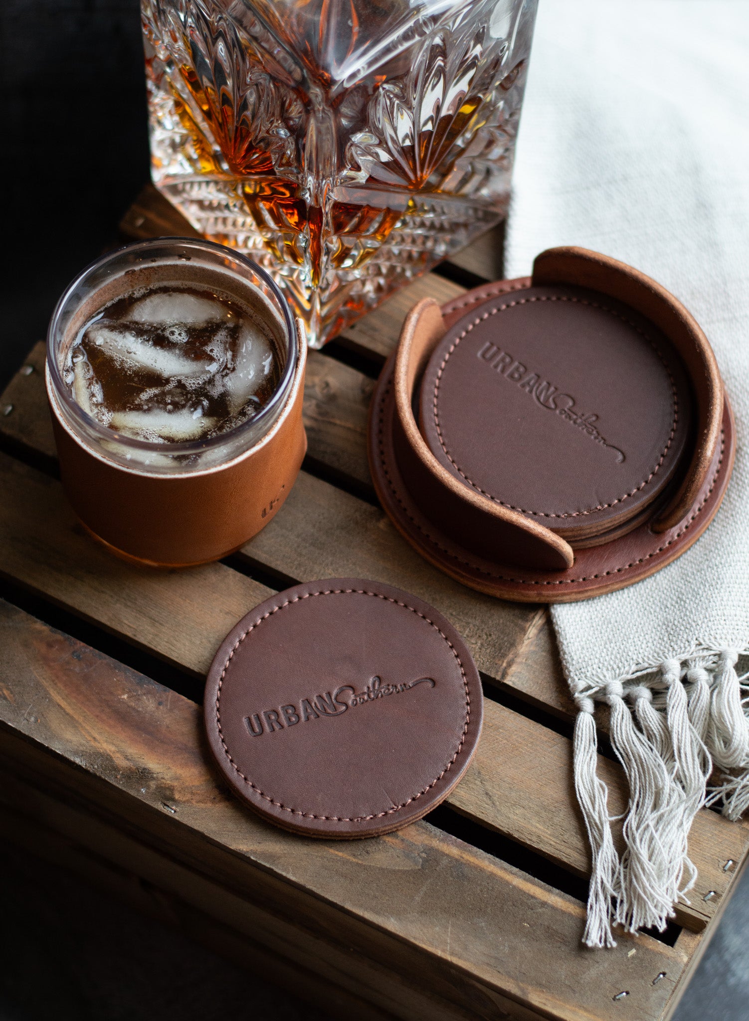 A set of Chestnut brown, Urban Southern branded coasters sit on top of a wooden crate with a glass of whiskey next to it.