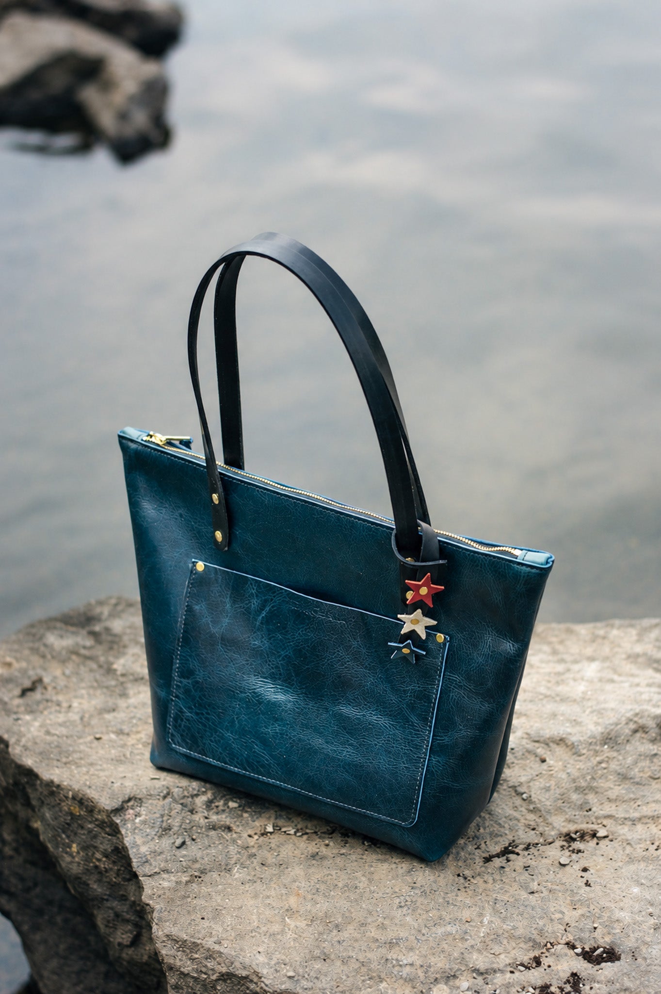 A blue leather tote with red white & blue heritage star bag charm looped around the handle sits on a rock near a body of water.