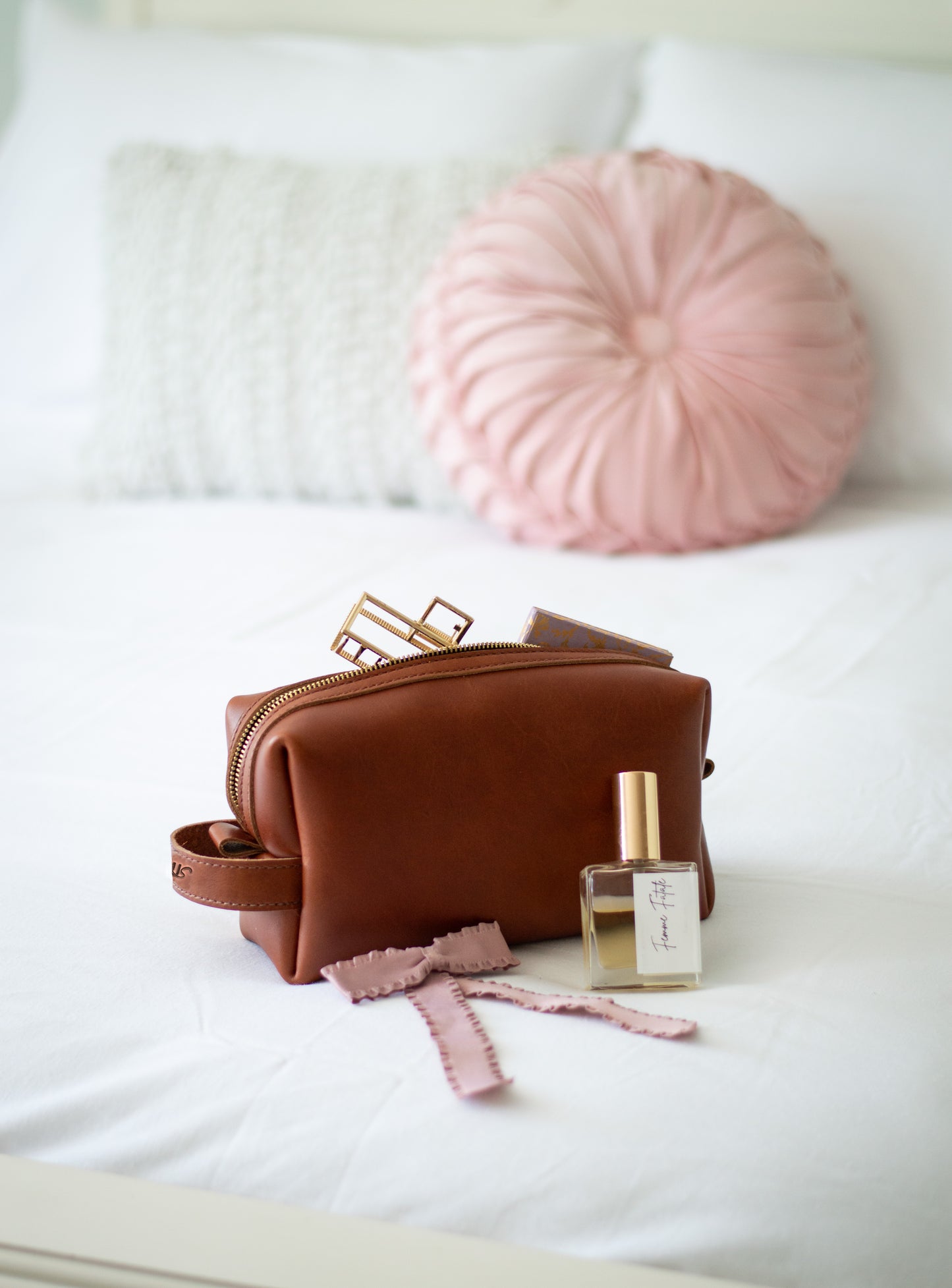 A leather travel dopp kit sits on a bed next to women's perfume & hair accessories.