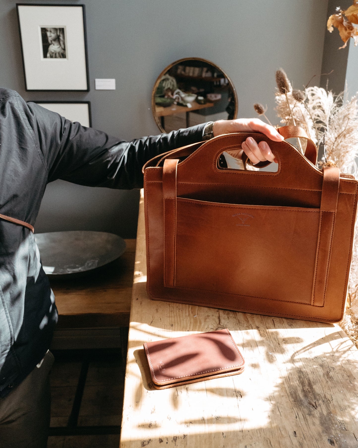 man holding a brown leather satchel by its grab handles on a wooden table with a neutral background