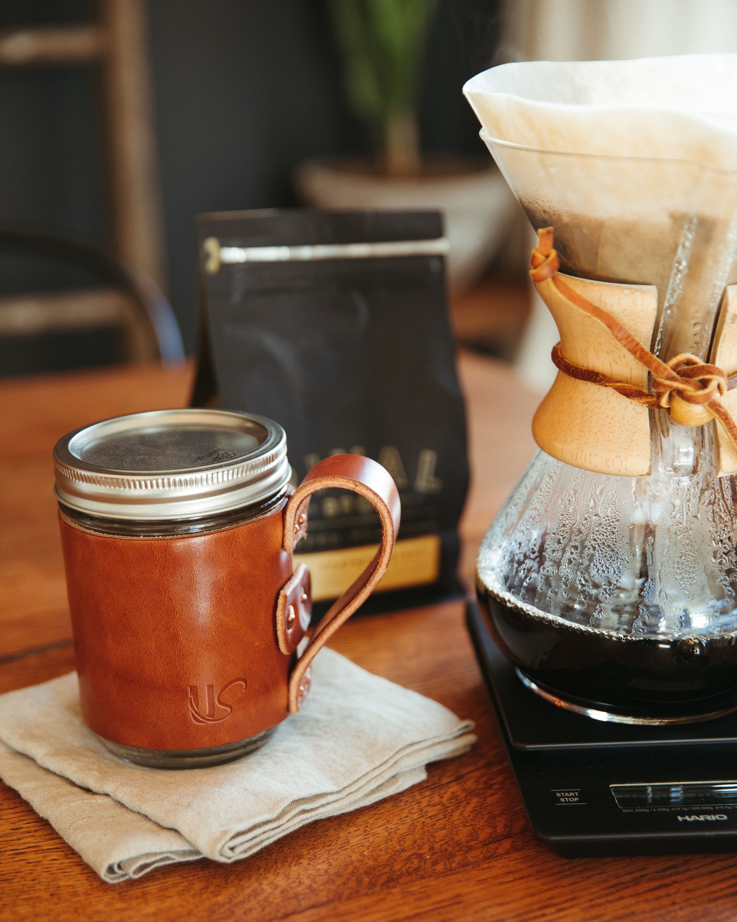 A beautiful, full-grain leather wrapped Mason Jar Mug, sits on a table next to a pot of coffee.