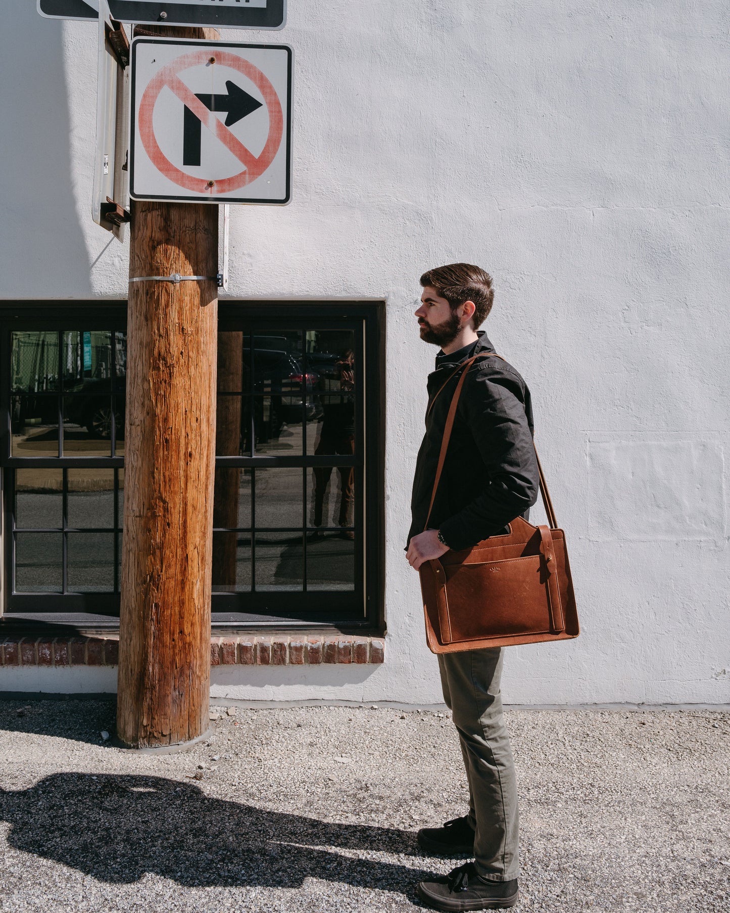 Man with a brown leather satchel on his shoulder standing next to a no right turn sign on a street.