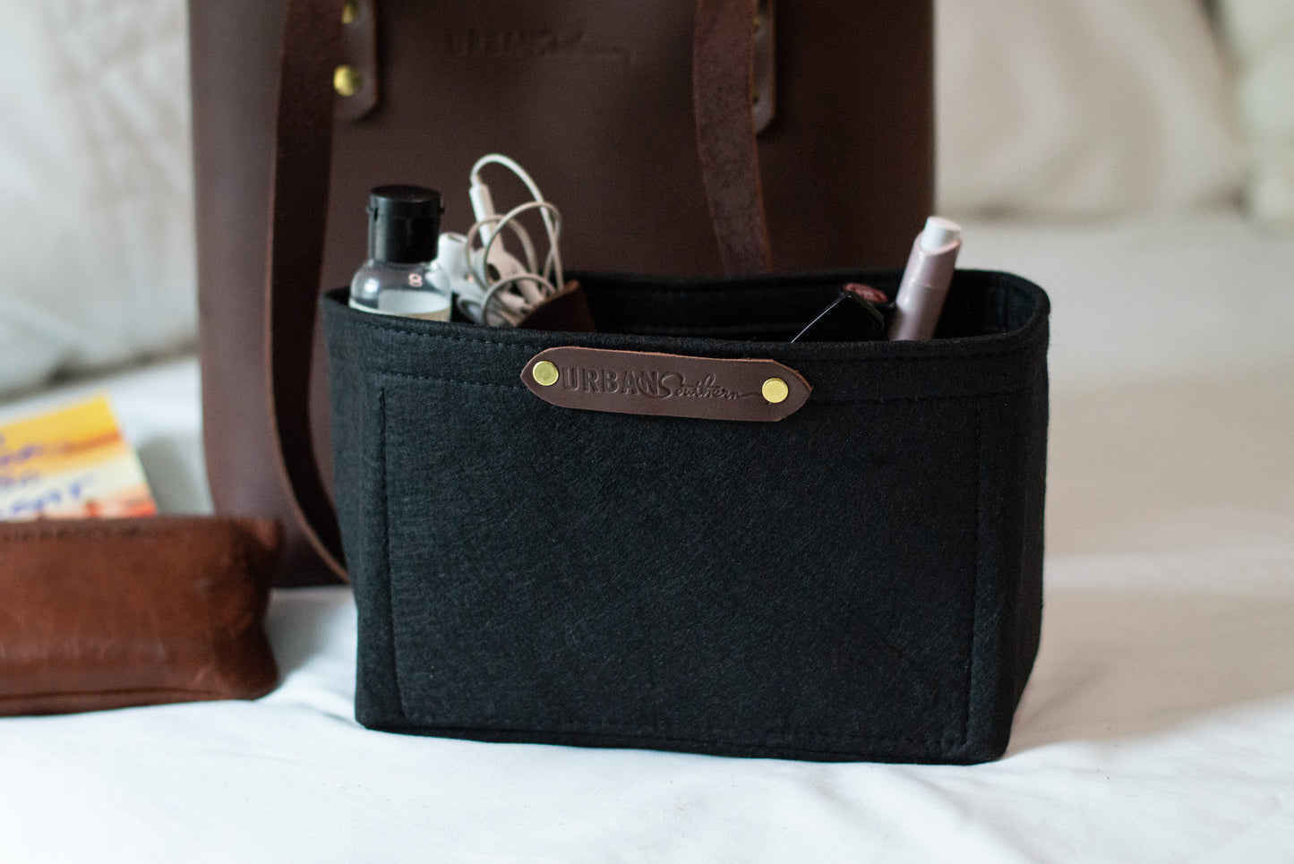 Sitting on a bed, a Mini Tote Insert is filled with everyday items, including lipstick, headphones etc.