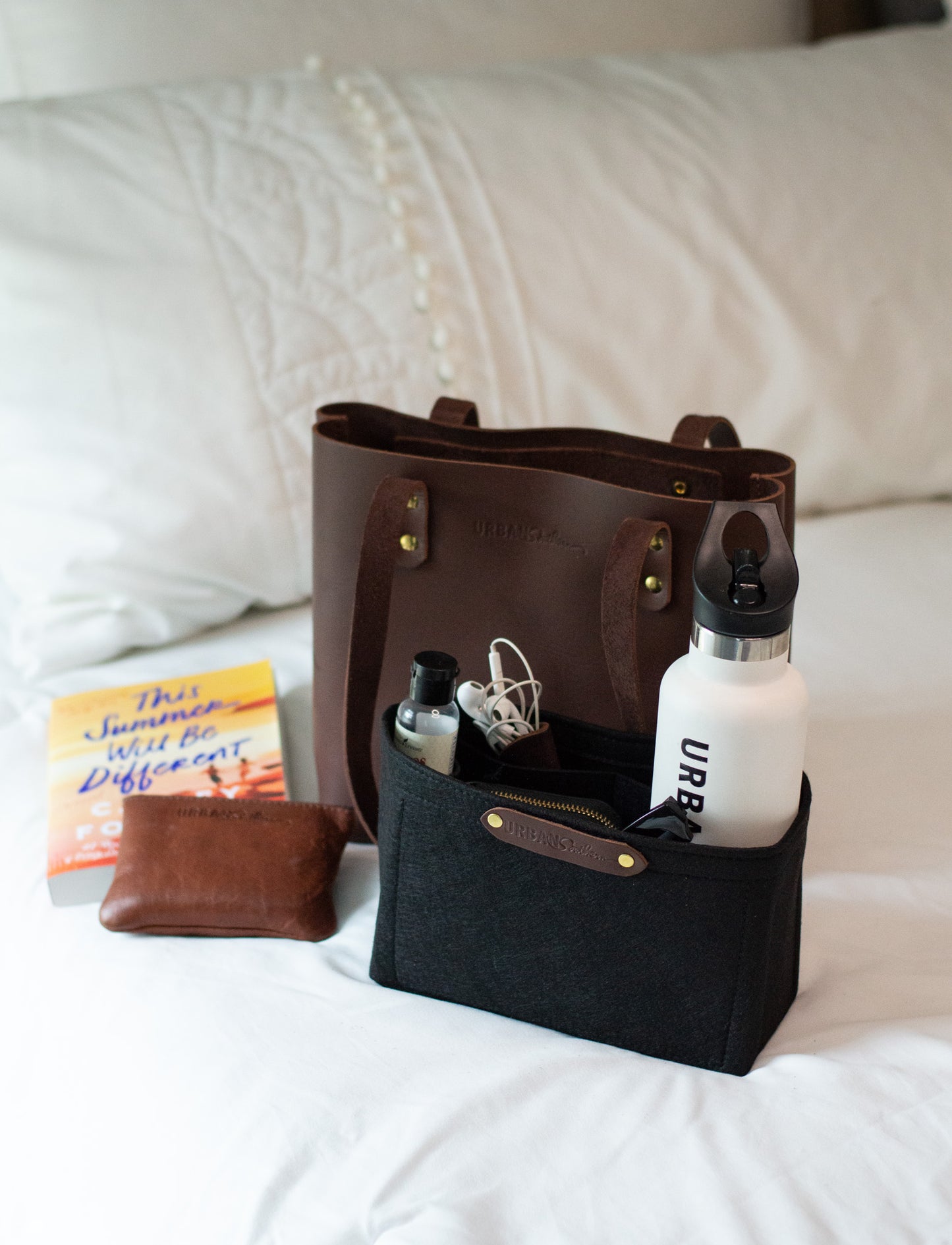Set out on a bed, a chestnut Mini Urban Tote sits next to a Mini Tote Insert filled with a variety of everyday items, including a water bottle, headphones, wallet etc.
