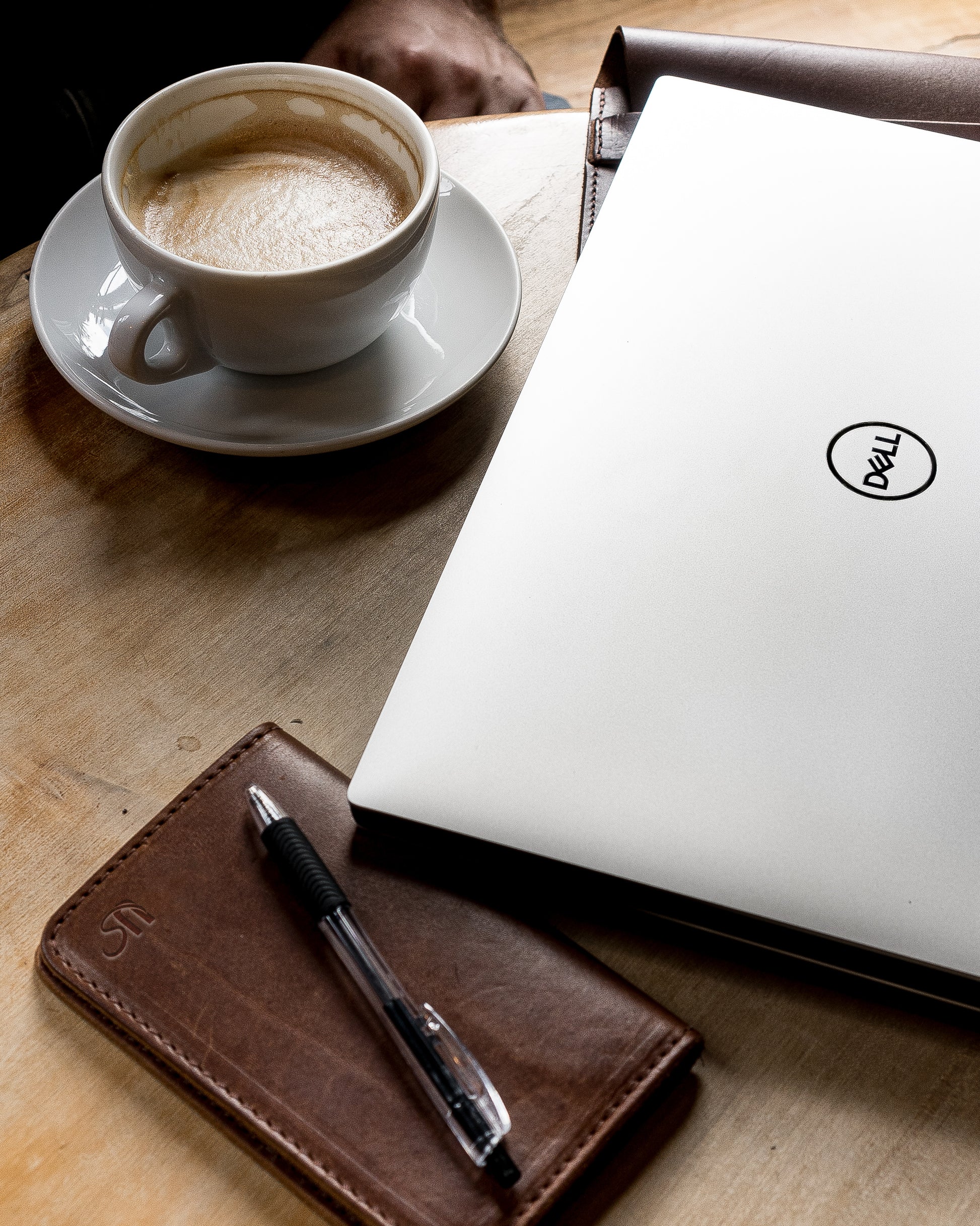 A leather notebook wallet sits on a cafe table next to a laptop and latte.