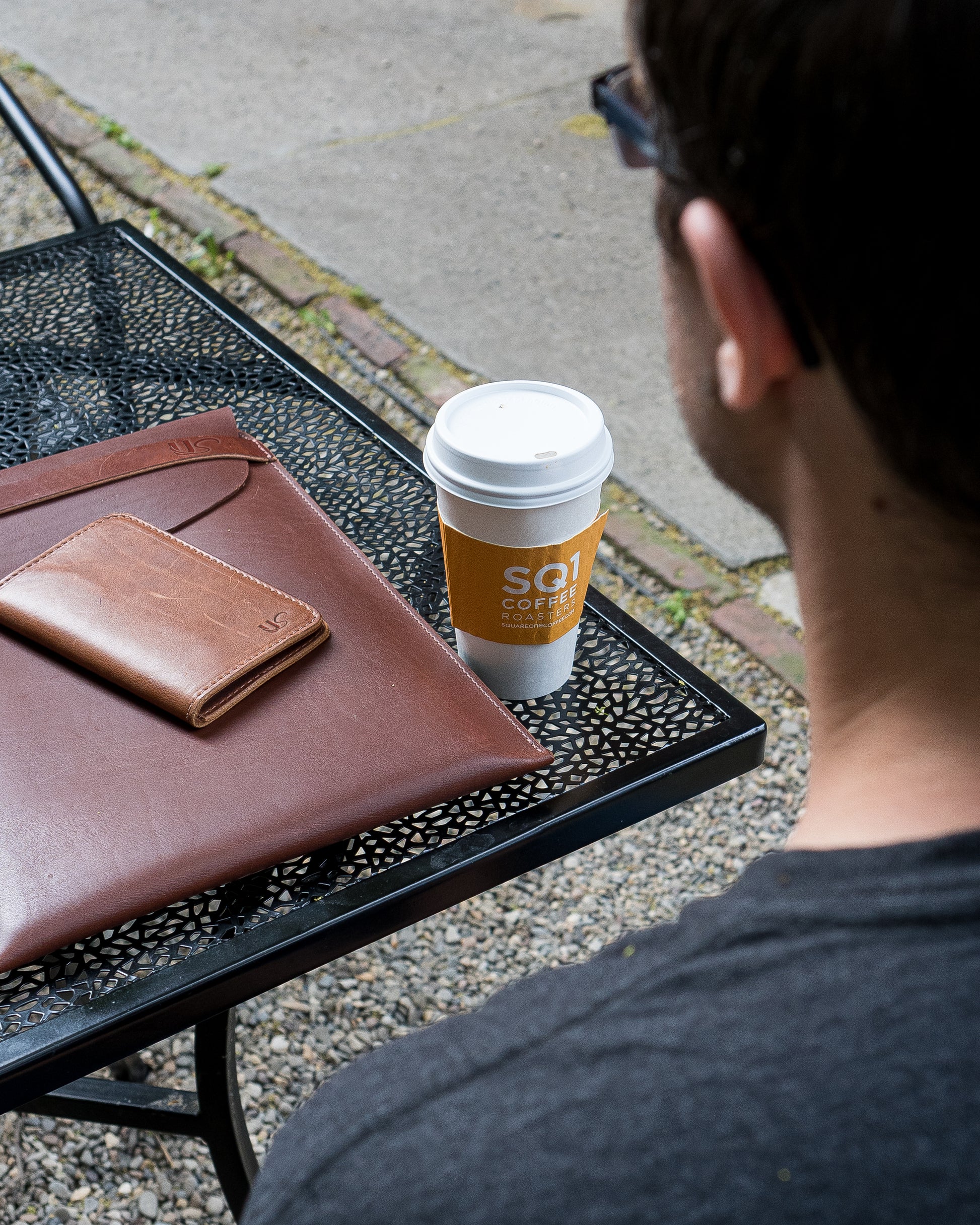 A man sitting at an outdoor cafe, enjoys his coffee, while a leather Notebook Wallet & Laptop Sleeve sit on the table next to him.
