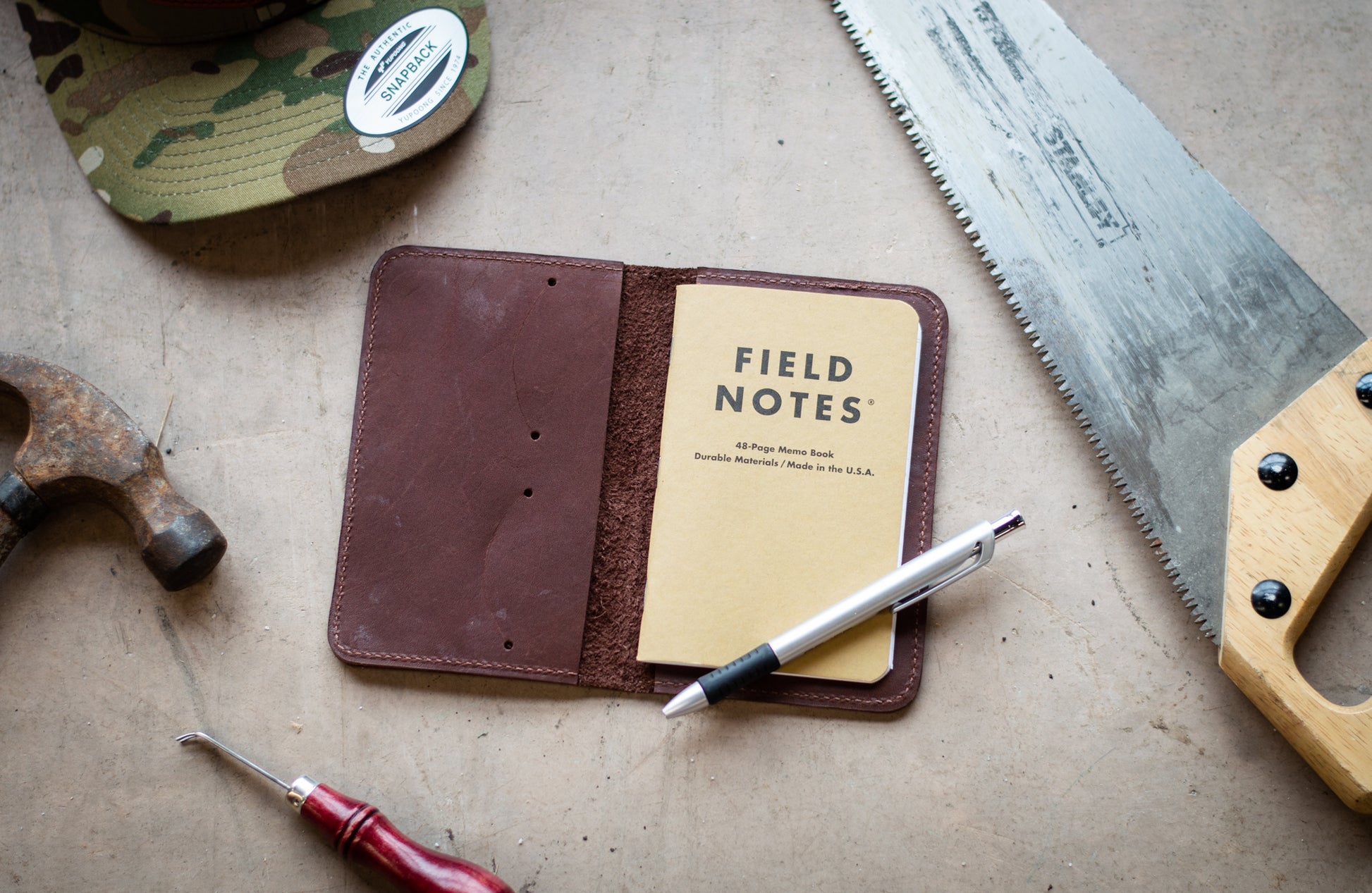 A leather Notebook Wallet, lays open next to a collection of tools. Making any day on the job site more organized with it's 48 page memo book & business card slots.