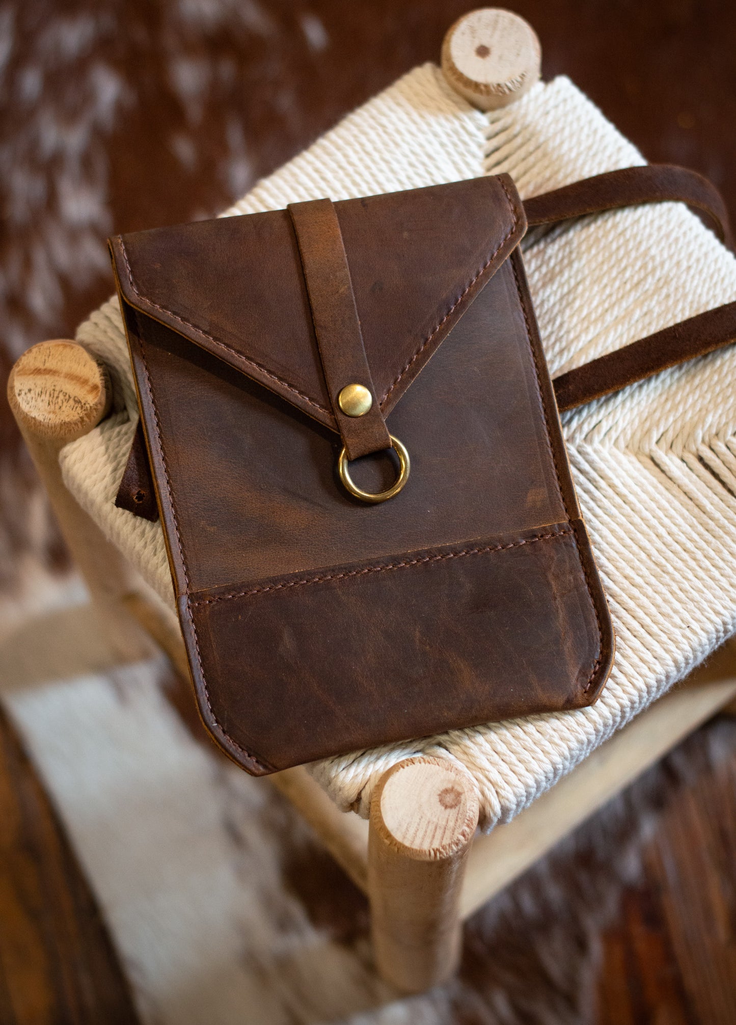 A Postal Crossbody in the color rustic brown, lays across a fabric stool.