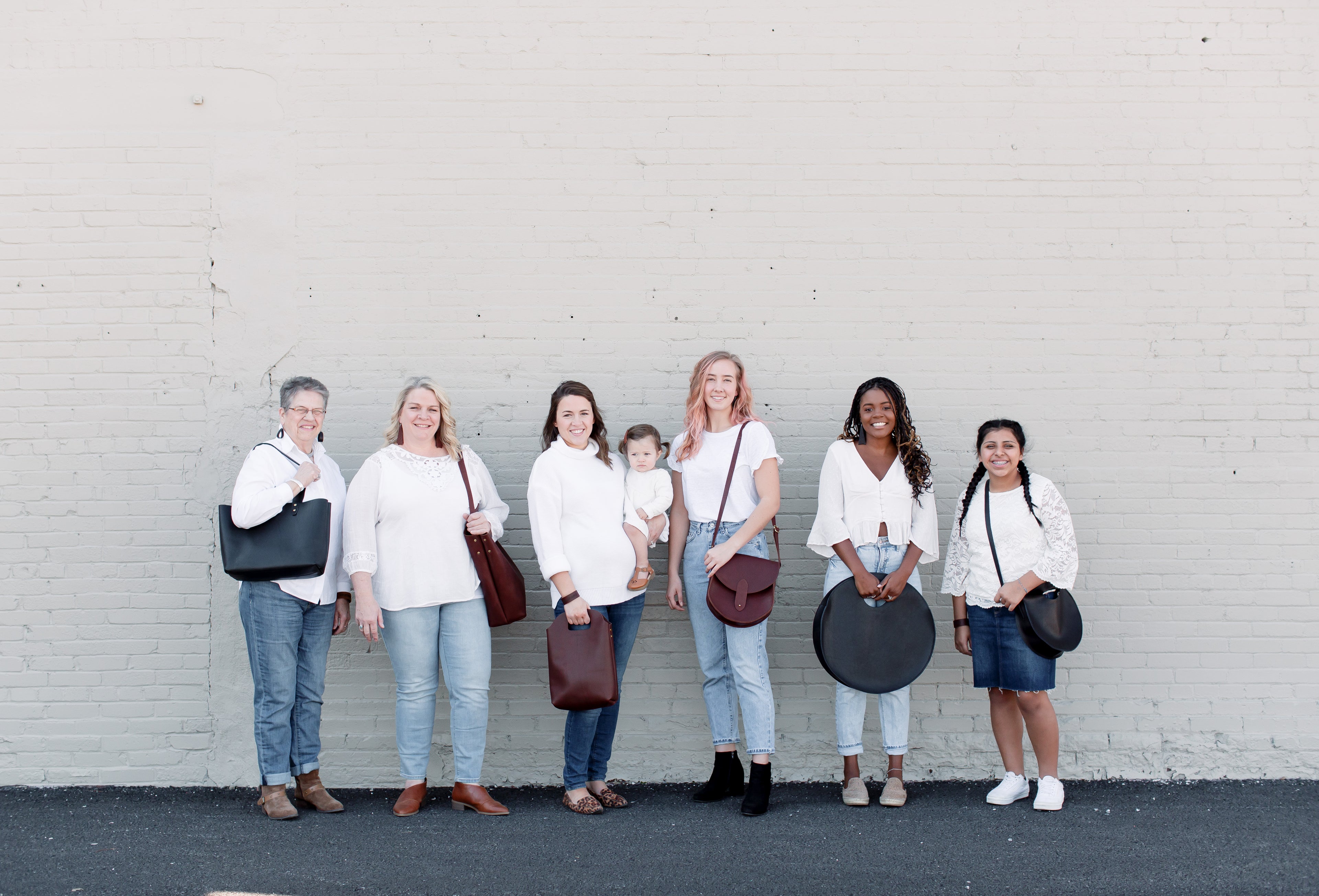 a group of women stand next to a white brick wall, each holding a leather handbag.