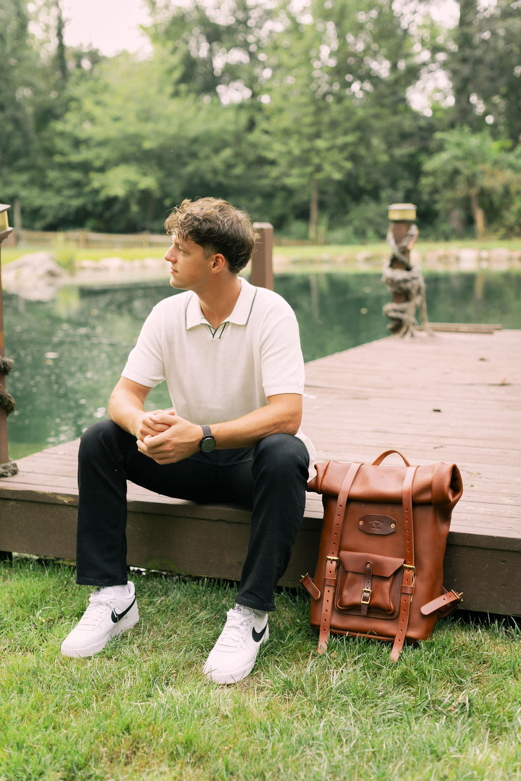 Man sitting on a dock with a brown leather backpack next to him, surrounded by greenery.