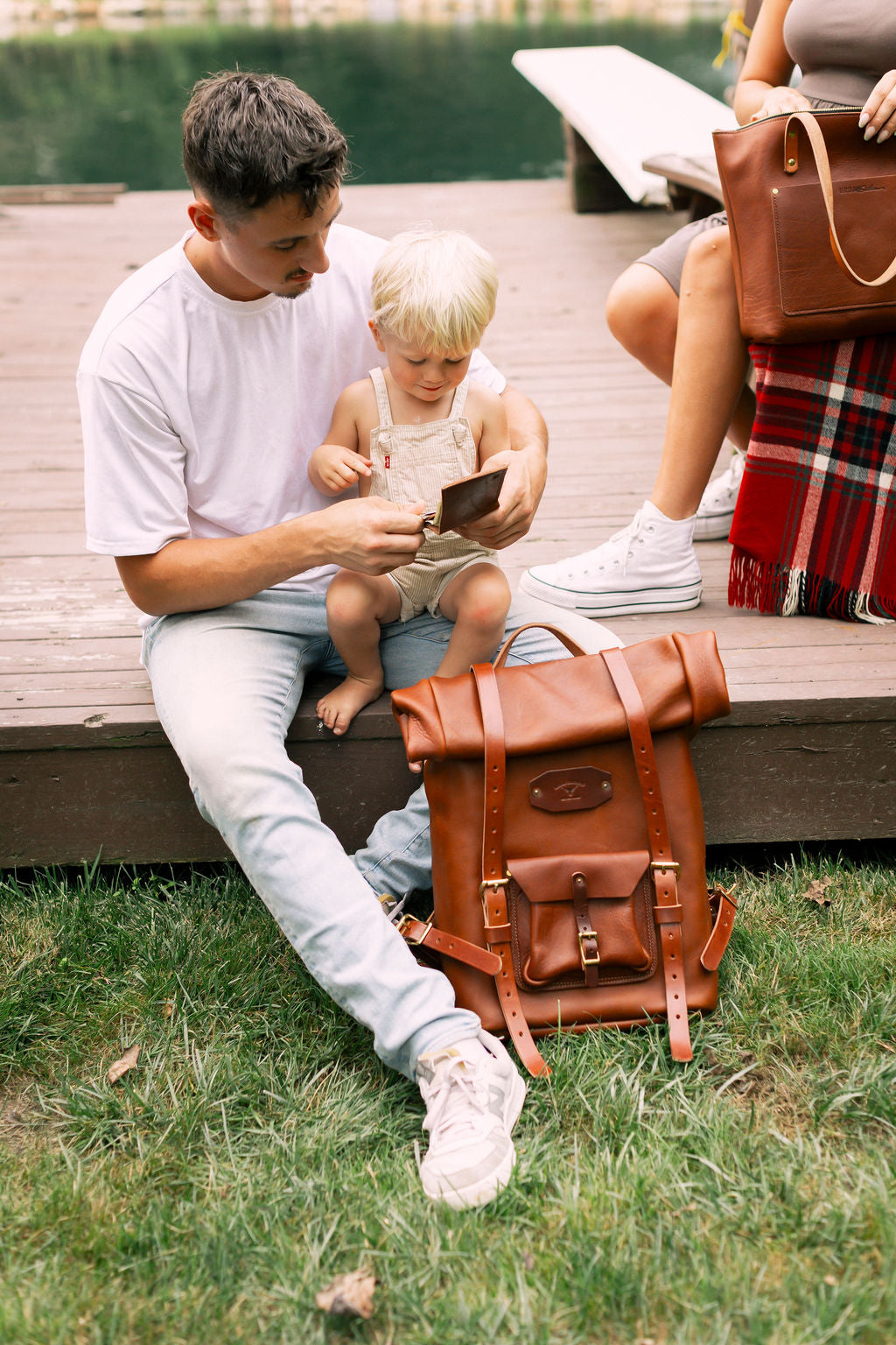 Man and child sitting on a wooden dock with a brown leather backpack nearby.