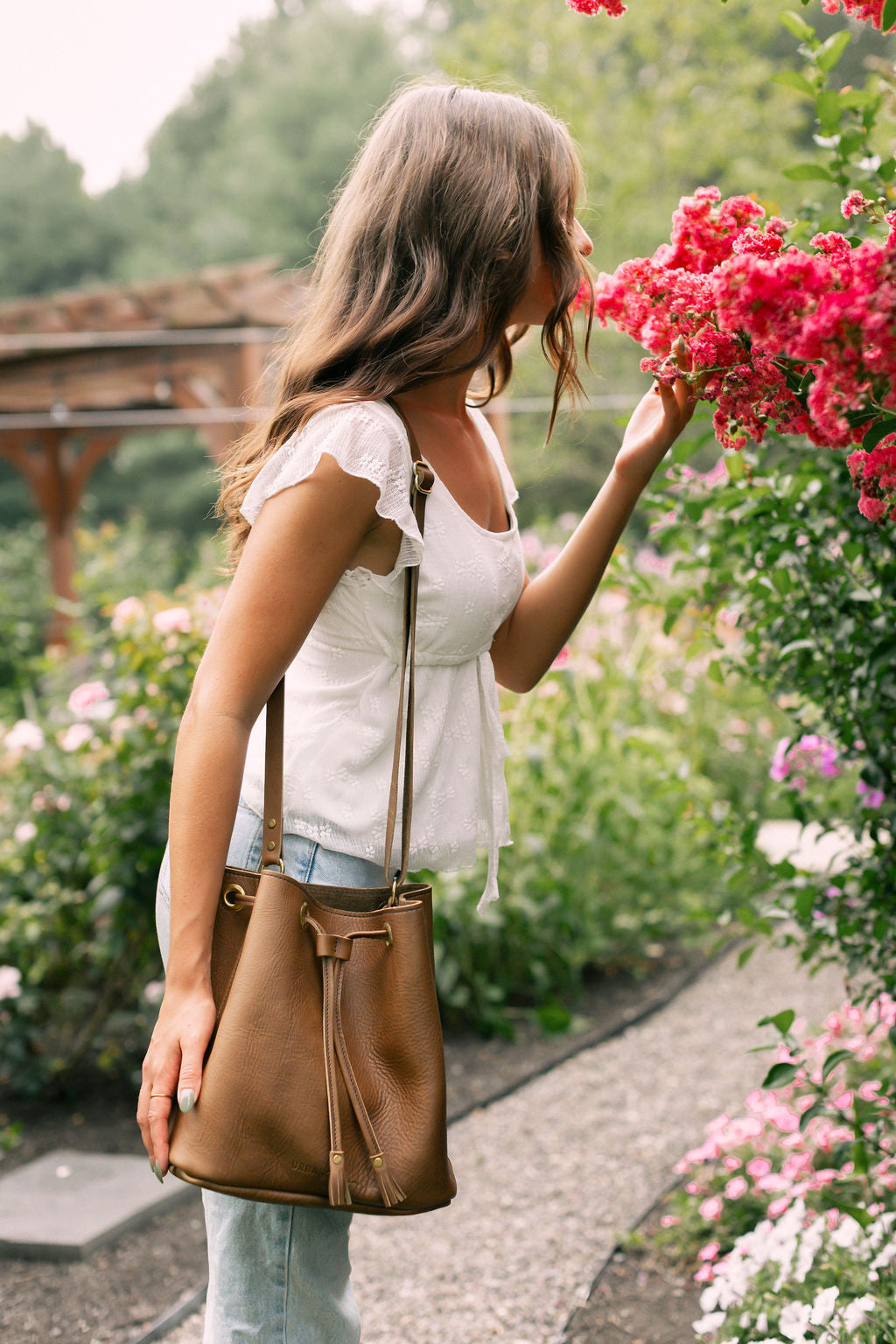 Woman holding a mocha leather bucket bag in a garden with pink flowers