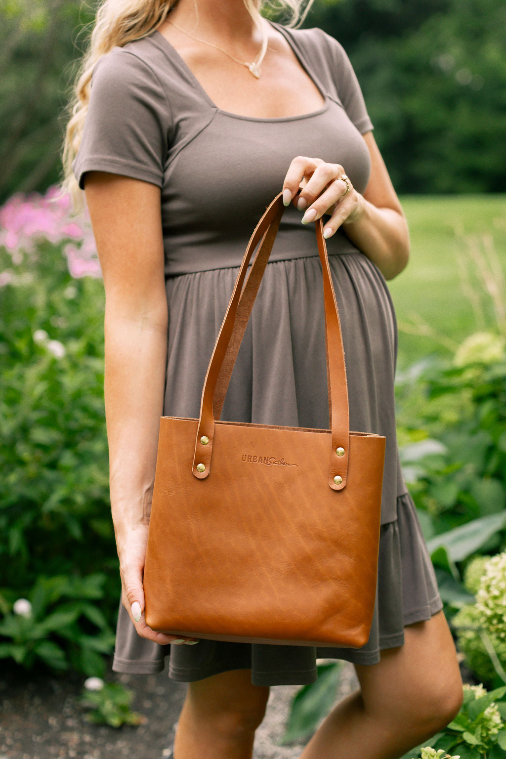 Woman holding a light brown leather tote bag outdoors with greenery in the background
