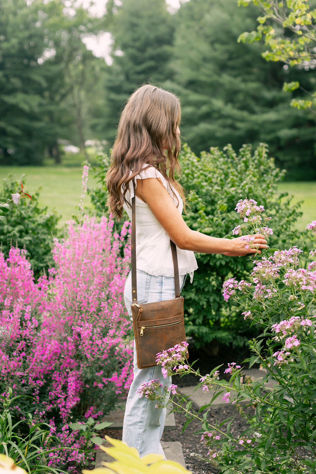 Woman standing in a garden with pink flowers and greenery, a rustic brown leather handbag hangs over her shoulder.