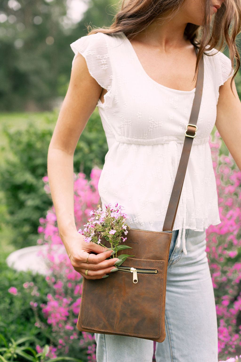 Woman holding a brown leather crossbody bag with pink flowers in a garden setting
