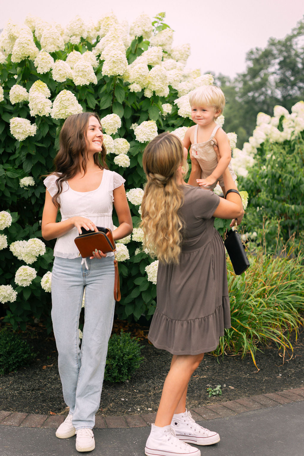 Two women holding leather zipper wallets stand in front of white flowers with a child