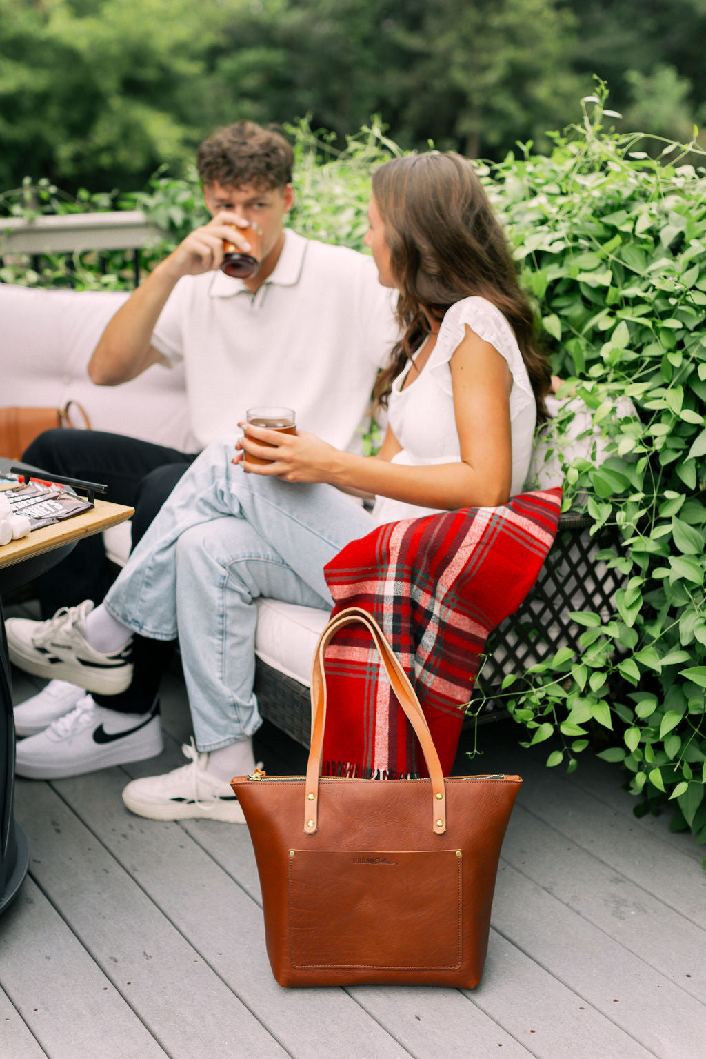 Man and woman sitting on a bench with a brown leather tote in front of them, surrounded by greenery.