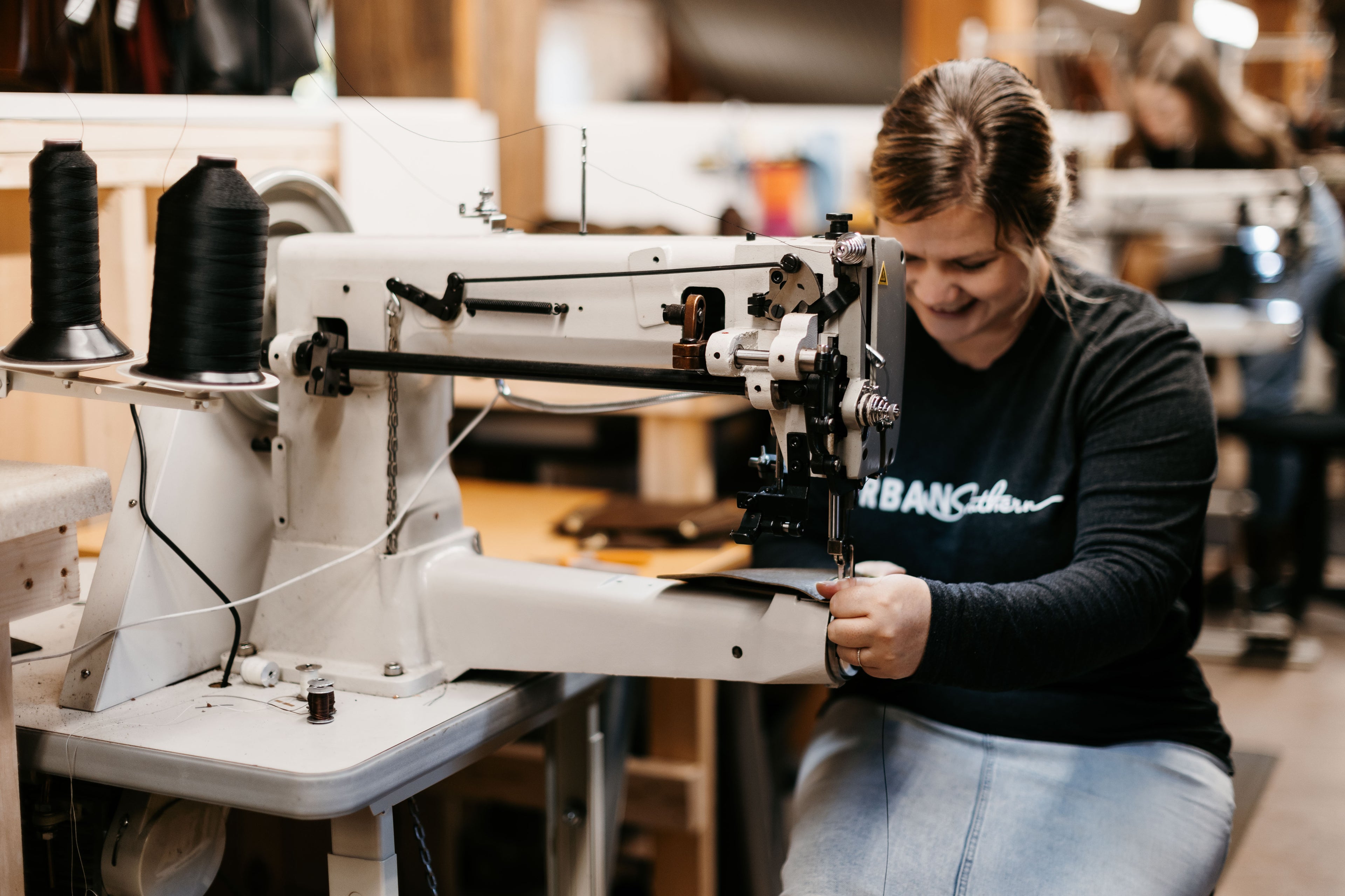 A woman sitting at a sewing machine, sews a leather handbag.