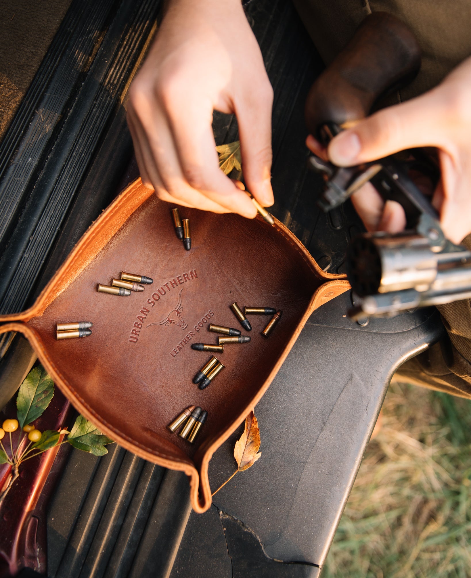 A leather valet tray filled with bullets, sits on the bed of a pickup truck, while a man loads a handgun, picking up a bullet from the this handy catch-all tray.