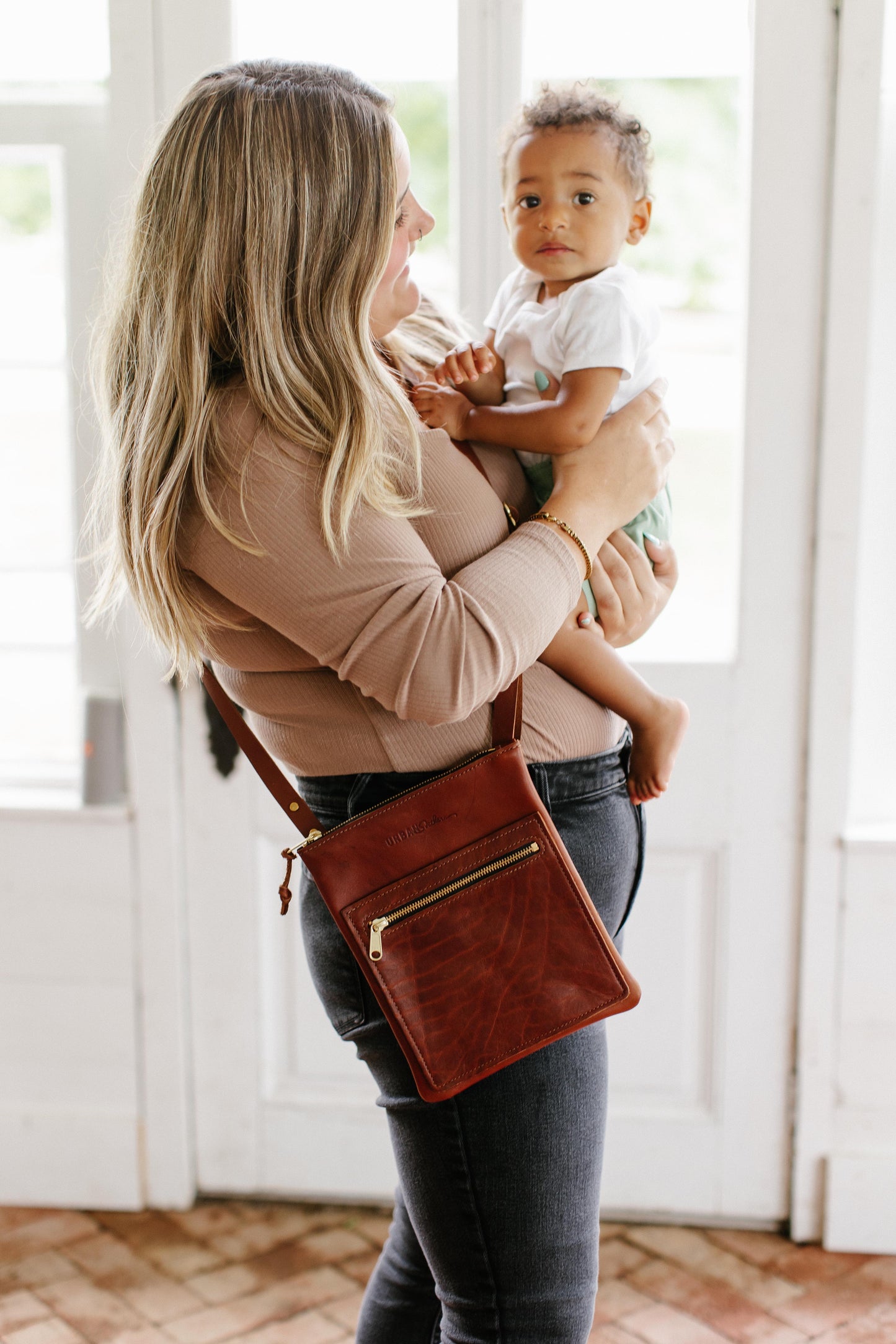 A woman holding a small child, wears a cognac Urban Sling Crossbody across her body.