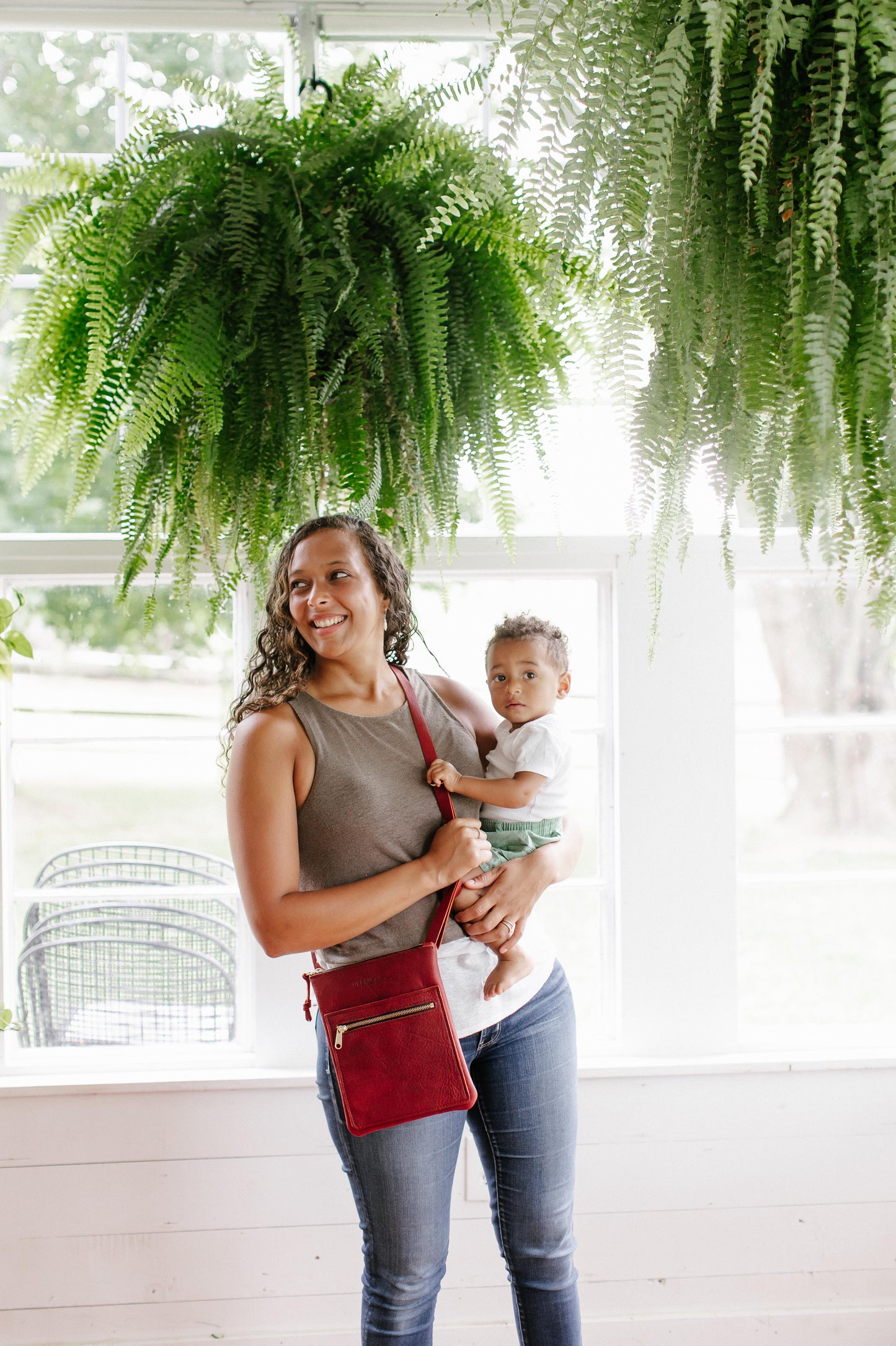 A Woman standing in a greenhouse, holds a small child in her arms while wearing a merlot Urban Slim Crossbody across her body.