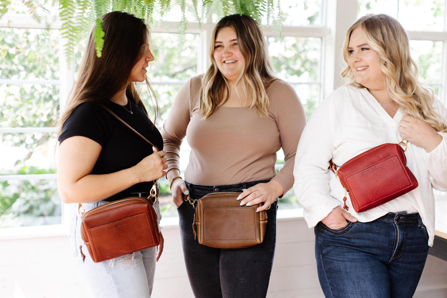 Three women stand in a greenhouse, each wearing an Urban Sling. Showcasing the variety of ways you can wear this bag.