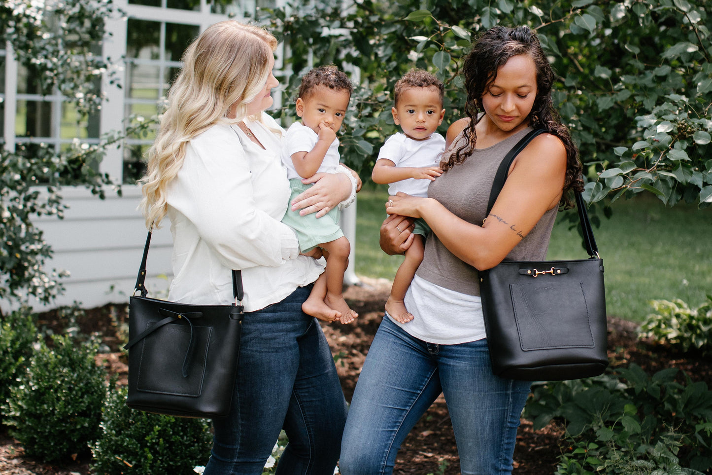 Two women stand in a garden, each holding a baby in their arms. One woman has a black Lexington Julep on her shoulder while the other woman has a black Lexington Bow on her shoulder.