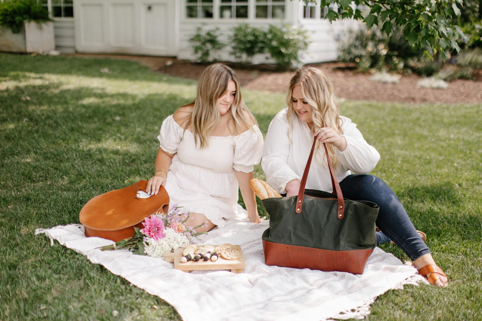 Two women picnic outside, one holding a Carrollton Circle tote in Honey while the other one holds a Large Canvas Tote in Hunter.