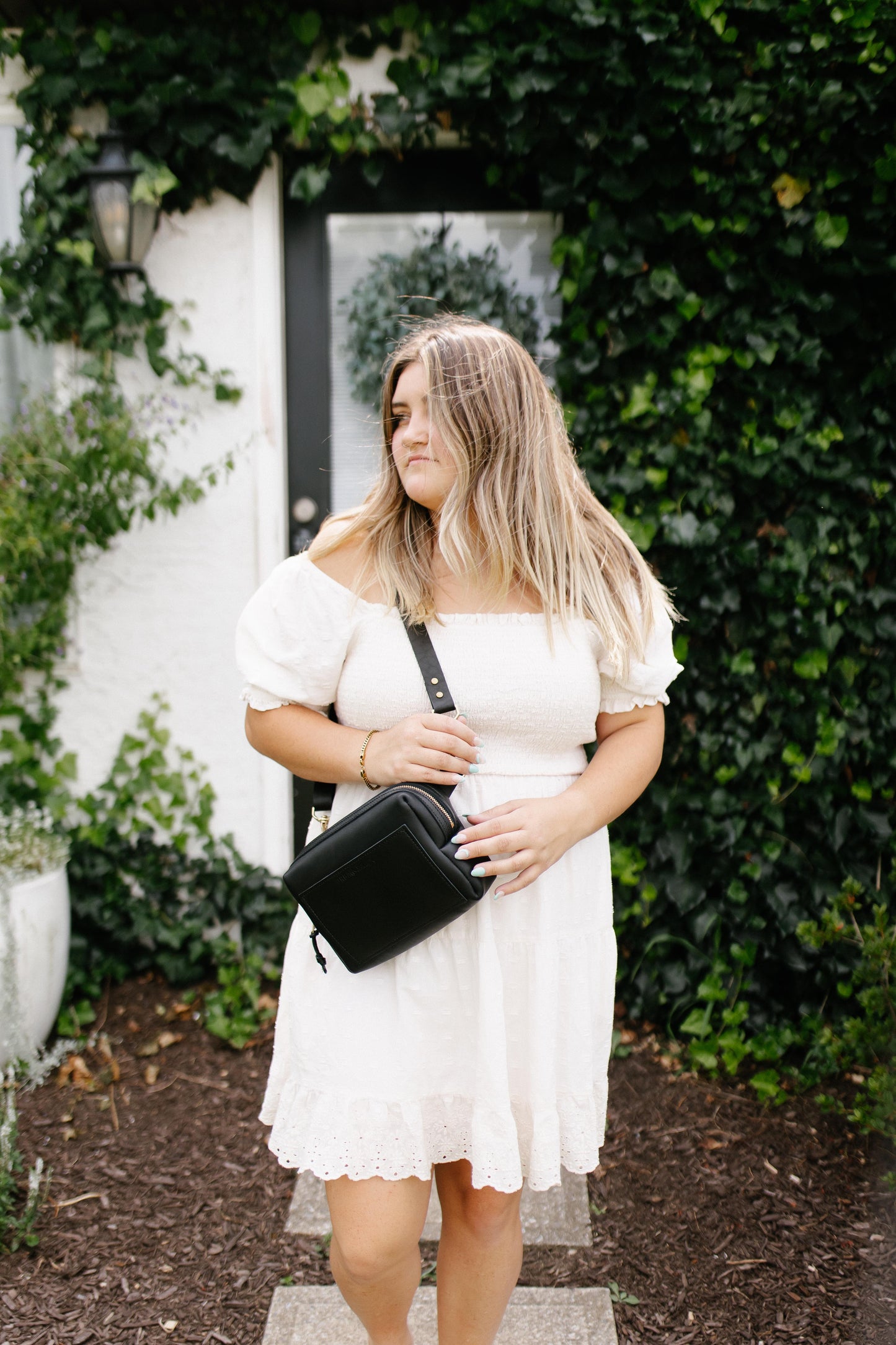 A woman standing in a garden, wears a black Urban Sling on her shoulder.