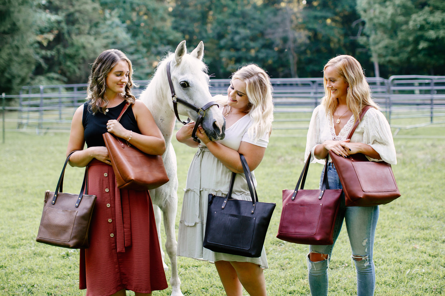 Three women stand next to a white pony in a pasture, each woman holding a variety of colors of the Urban Zipper Tote.