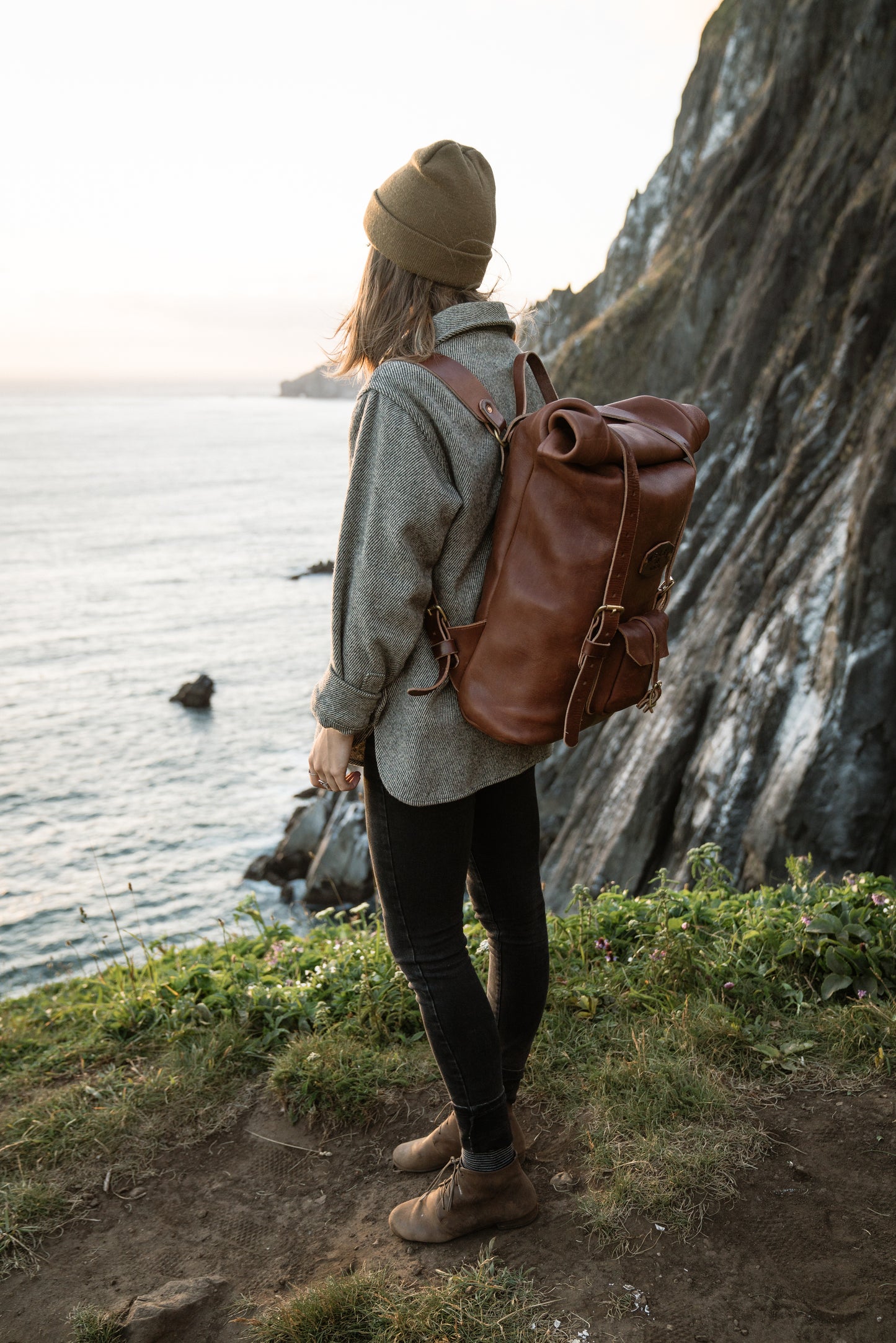 A woman wears a brown backpack standing by a body of water and rocky cliffs.