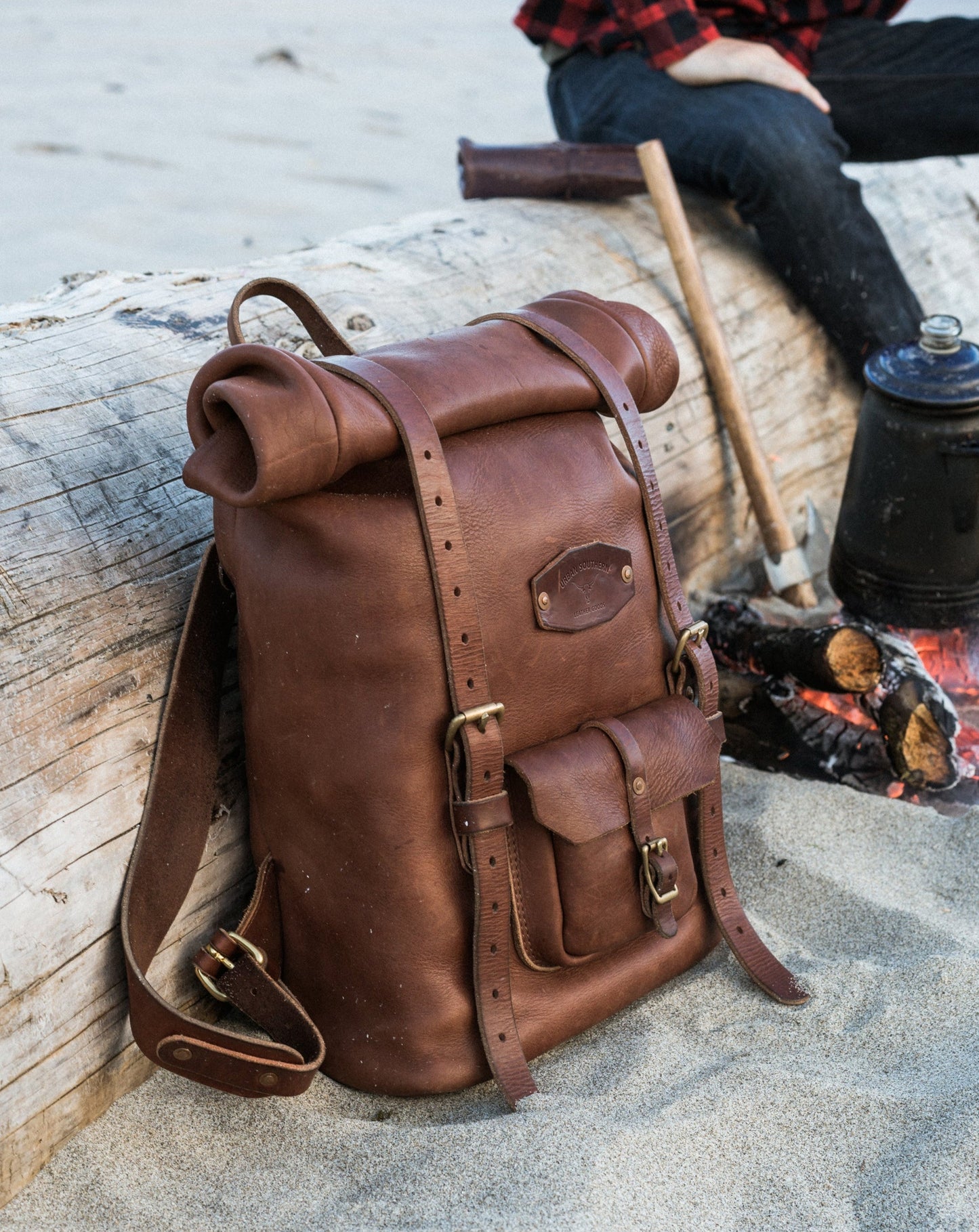 Brown leather backpack on a log with a person sitting nearby at a campfire.