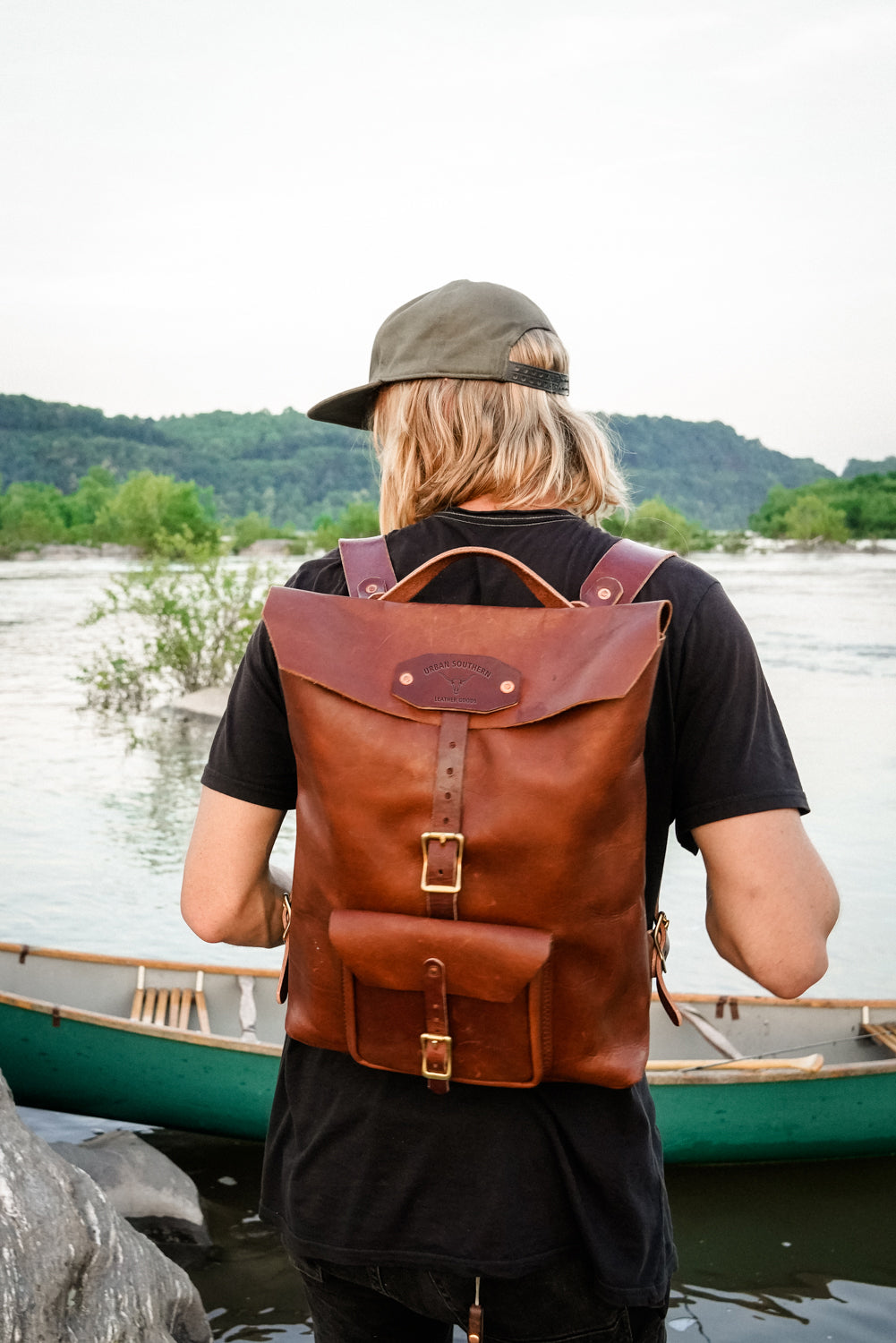 Person wearing a brown leather backpack by a lake with a canoe in the background