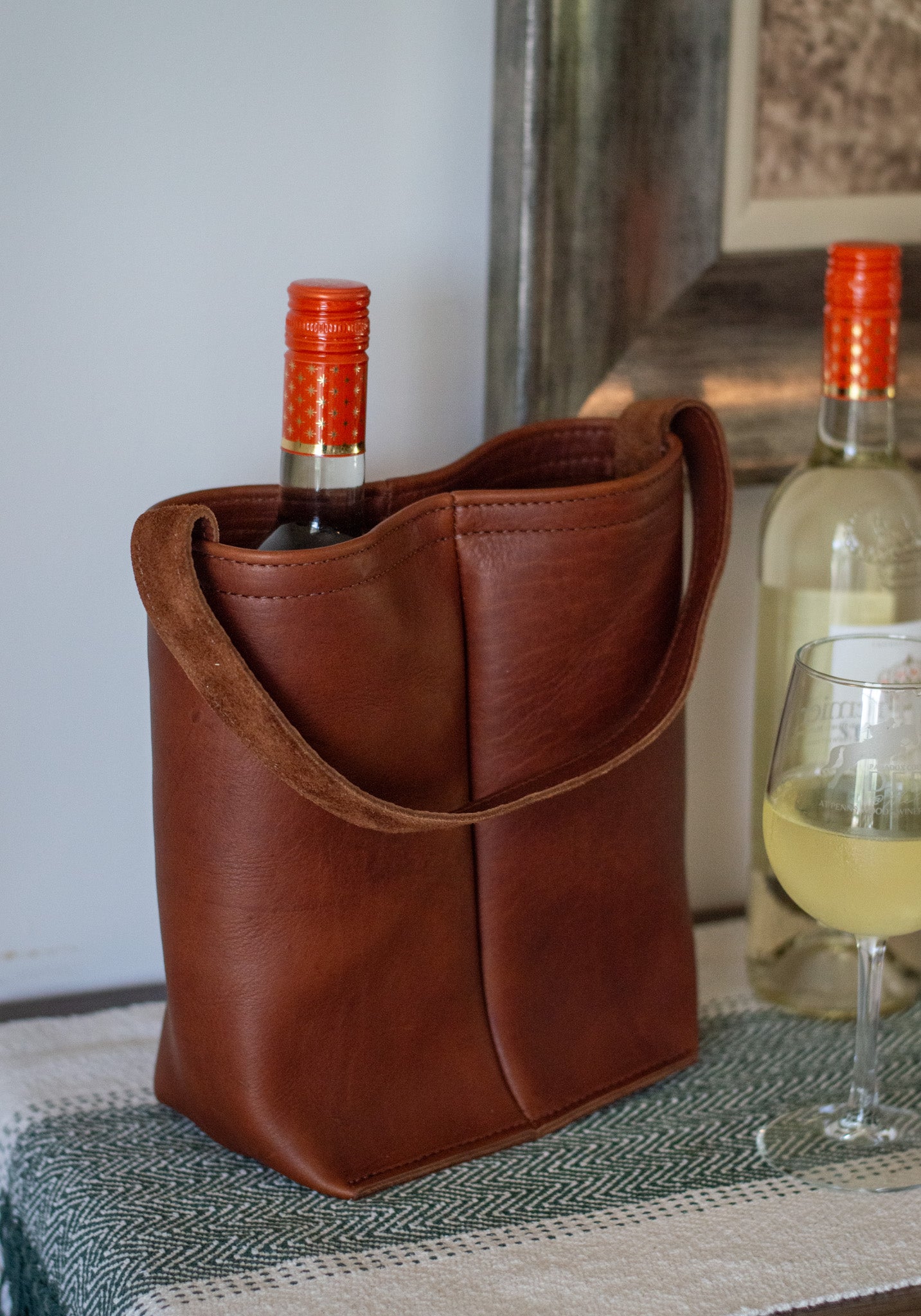 Leather wine tote, carrying a bottle of wine, sitting on a table with a glass of wine next to it.