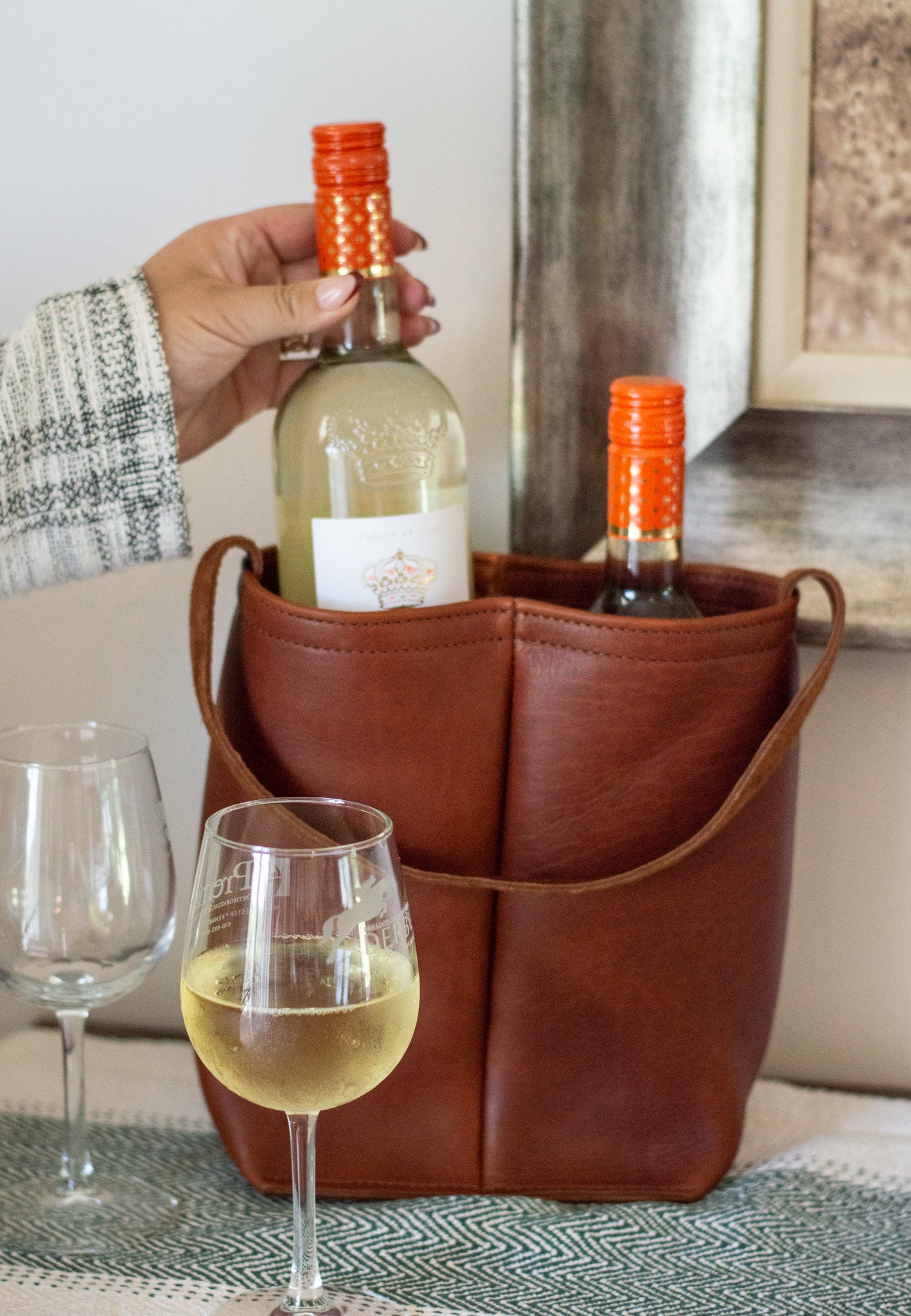 A Cognac leather Dual Wine Tote, sits on a table filled with two bottles of wine. A woman reaches for one of the bottles.