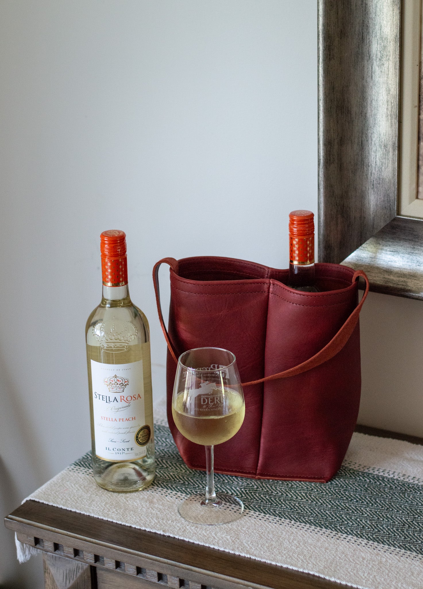 A merlot Double Wine Tote filled with a bottle of wine, sits next to a glass of wine & an additional bottle of white wine.