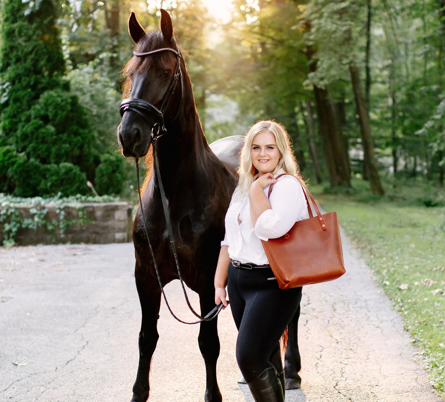 A woman holds the reins of a bridle on a black horse, while wearing an Urban Classic Tote on her shoulder.