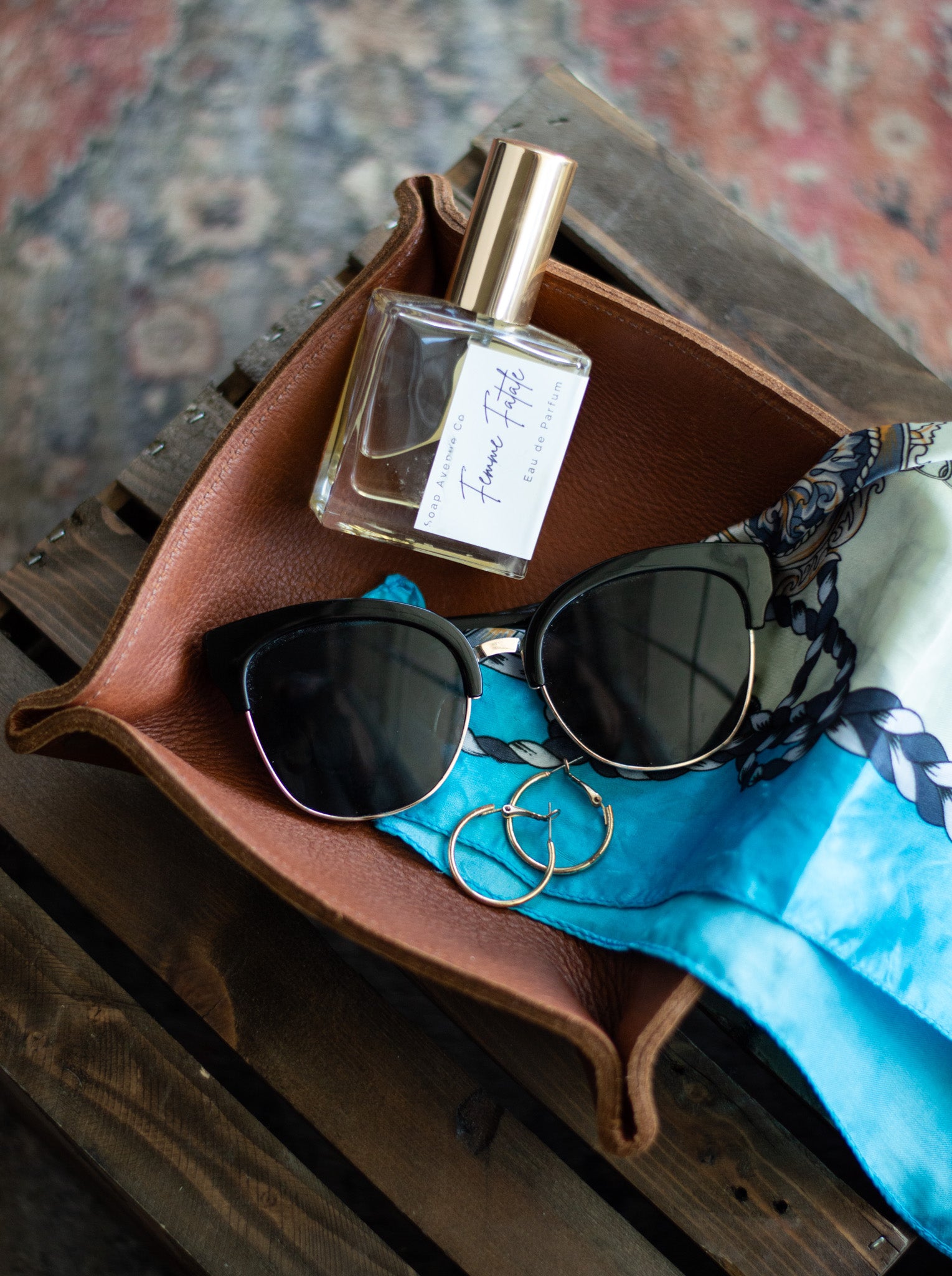 A leather valet tray is filled with women's perfume, sunglasses, earrings & a scarf.