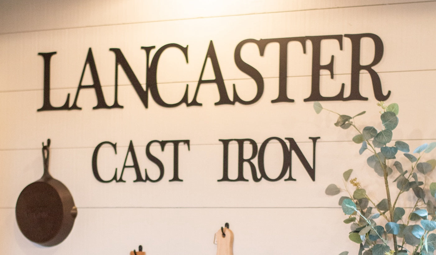 A white wall with a metal "Lancaster Cast Iron" sign hanging alongside a cast iron skillet.
