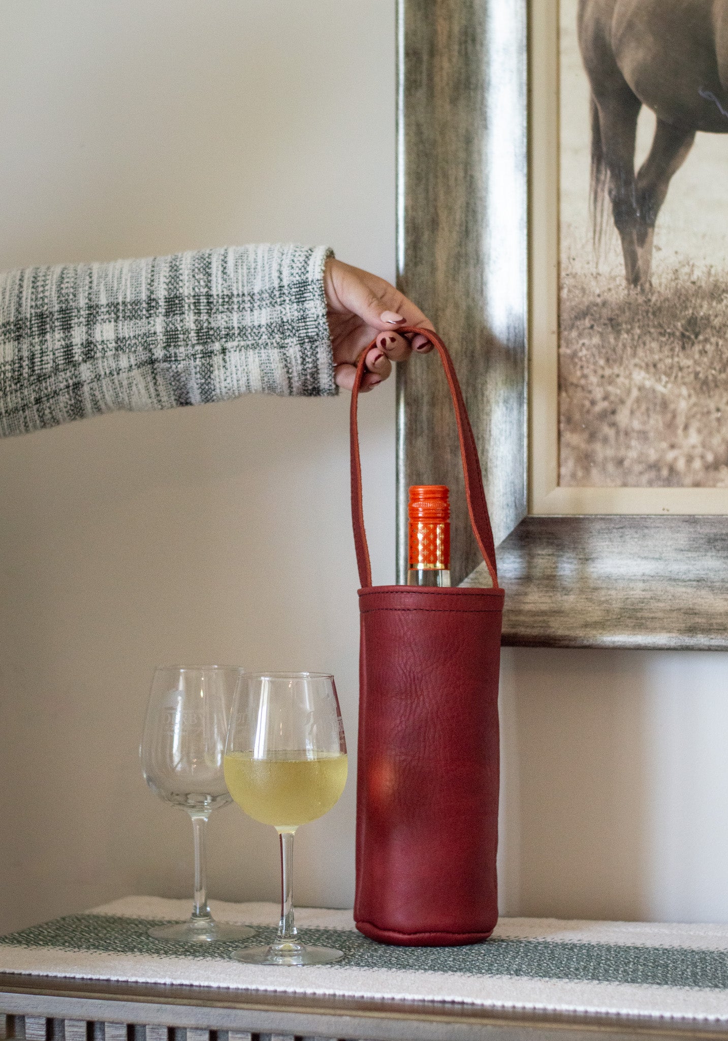 A woman holds the handle to a merlot Urban Wine Tote filled with a bottle of wine. Sitting next to the leather tote are two glasses of wine.