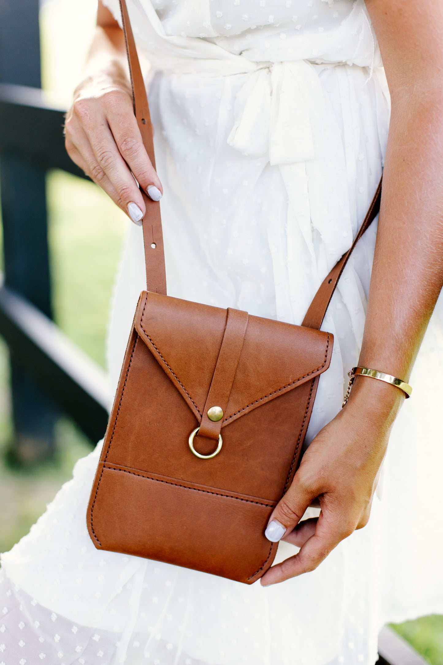 A woman holds a honey Postal Crossbody, showcasing the brass ring detail on the front of the leather handbag.